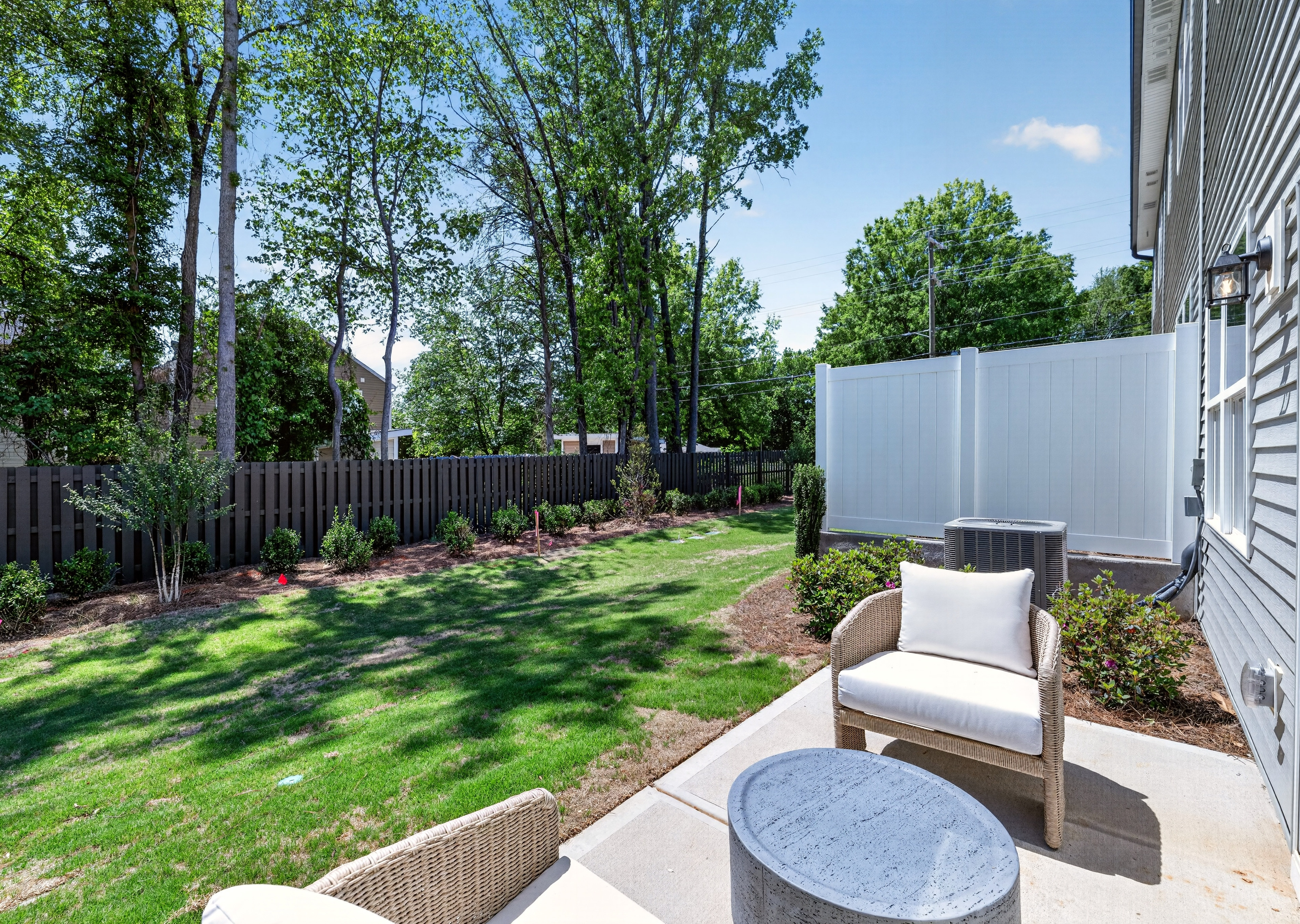 A patio with outdoor furniture sits on a concrete slab in the foreground, while a lush green lawn and trees fill the background.