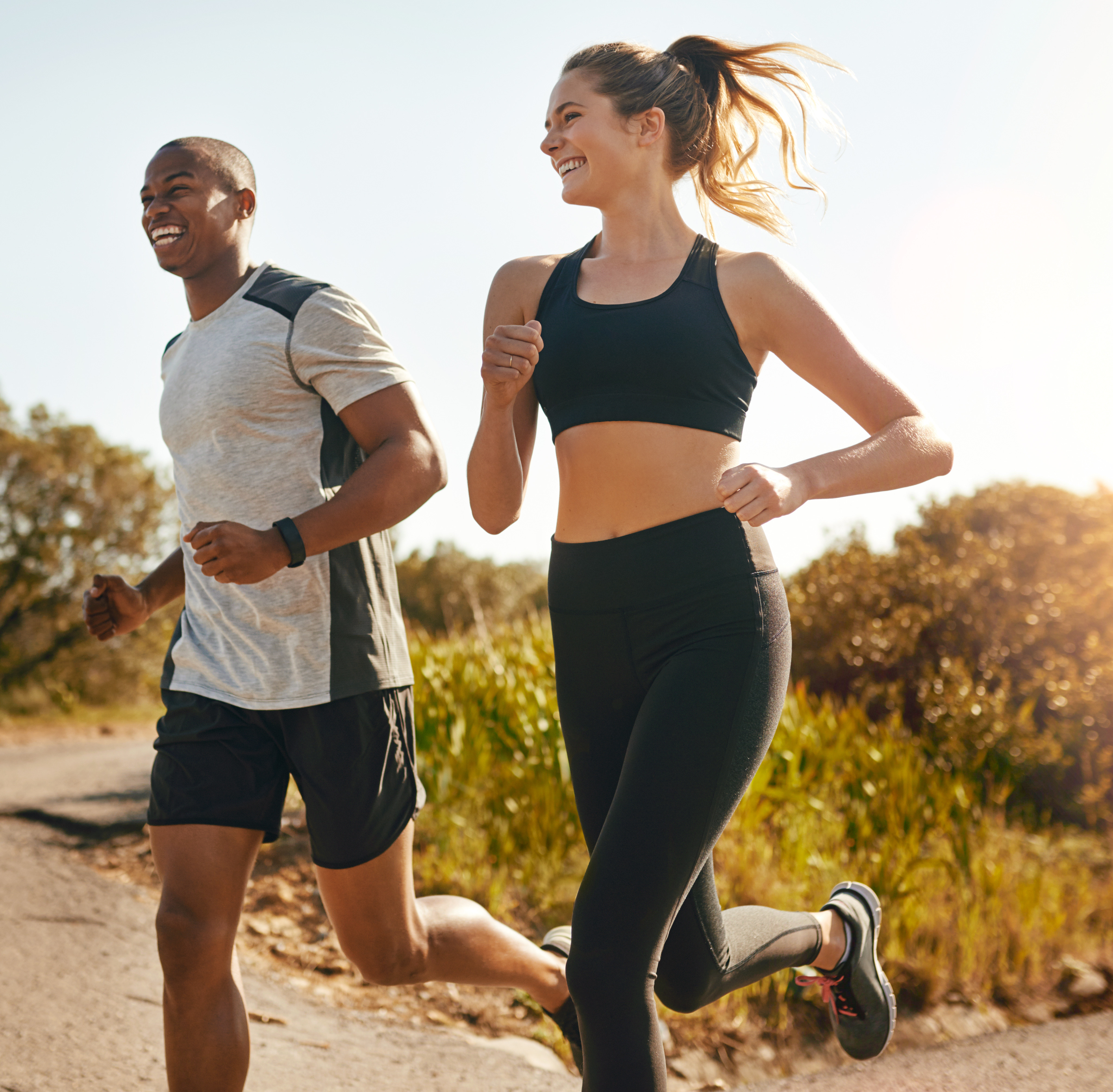 Two people, a man and a woman, are jogging together on a dirt path surrounded by lush greenery and sunlight filtering through the trees.
