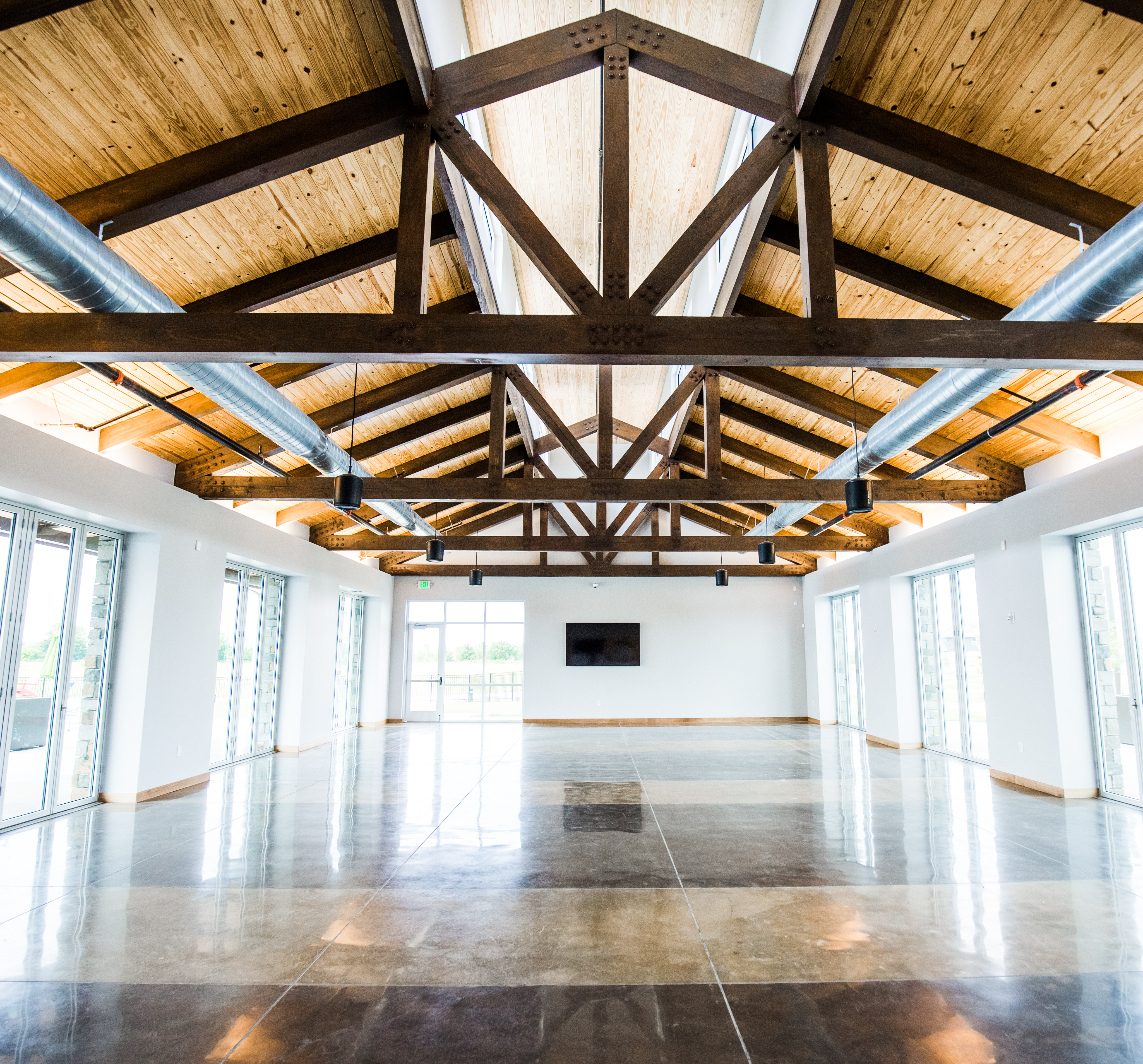 A spacious and well-lit interior with a high, wooden-beamed ceiling, polished hardwood floors, and large windows allowing natural light to flood the room.