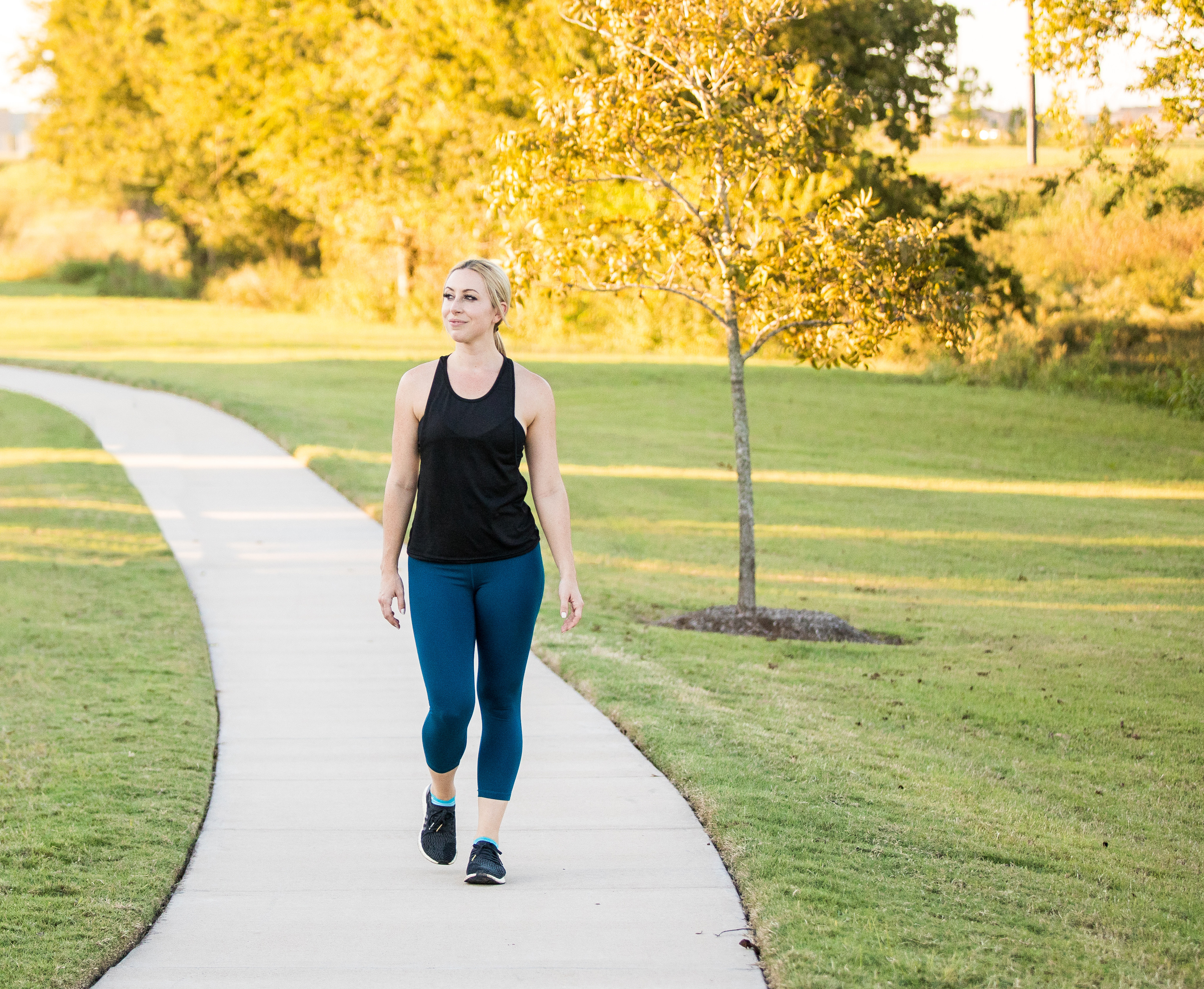 A woman in athletic clothing walks along a paved path surrounded by lush greenery and vibrant yellow trees in a park setting.