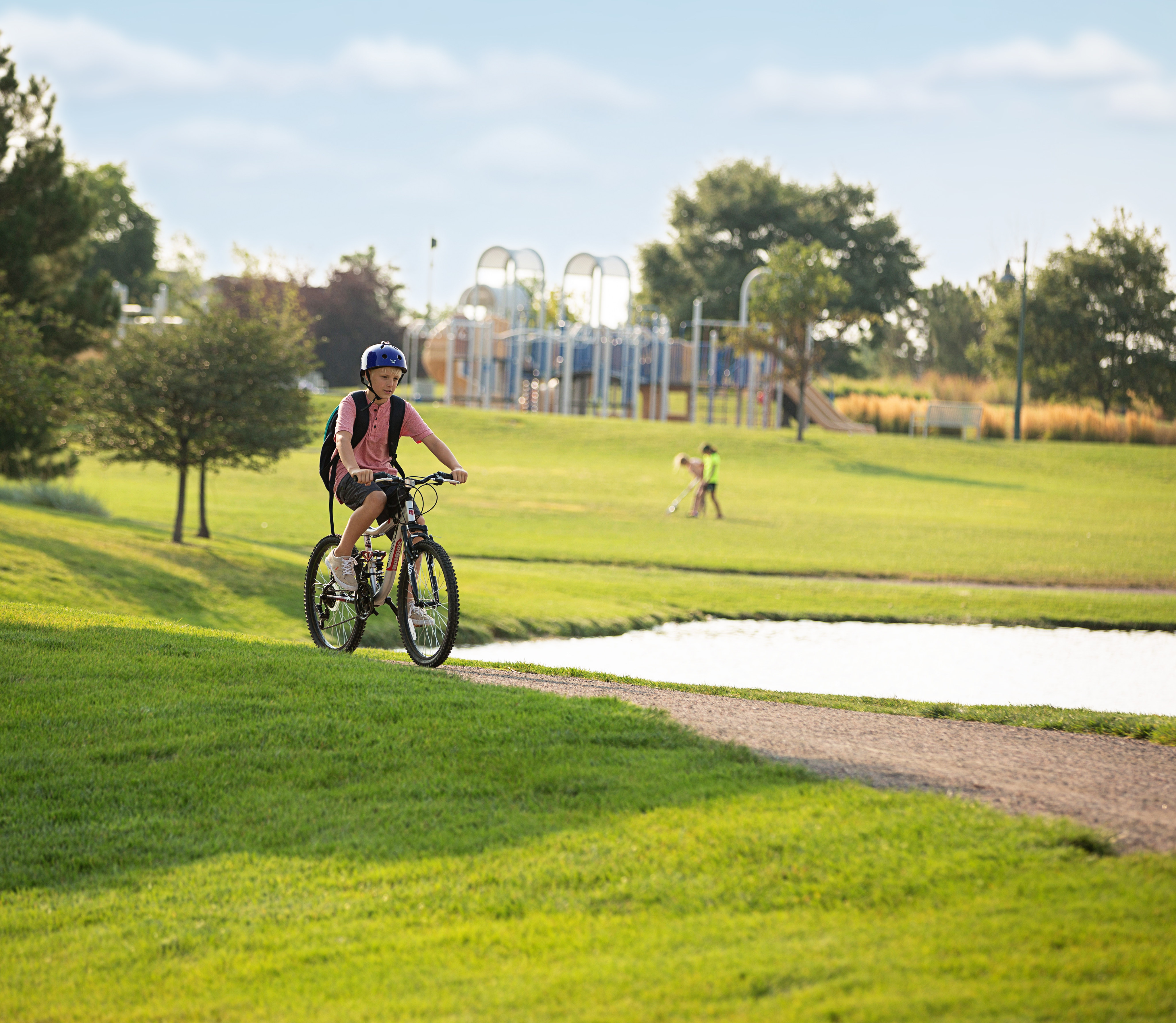 A person on a bicycle rides along a paved path through a lush, green park with trees and playground equipment visible in the background.