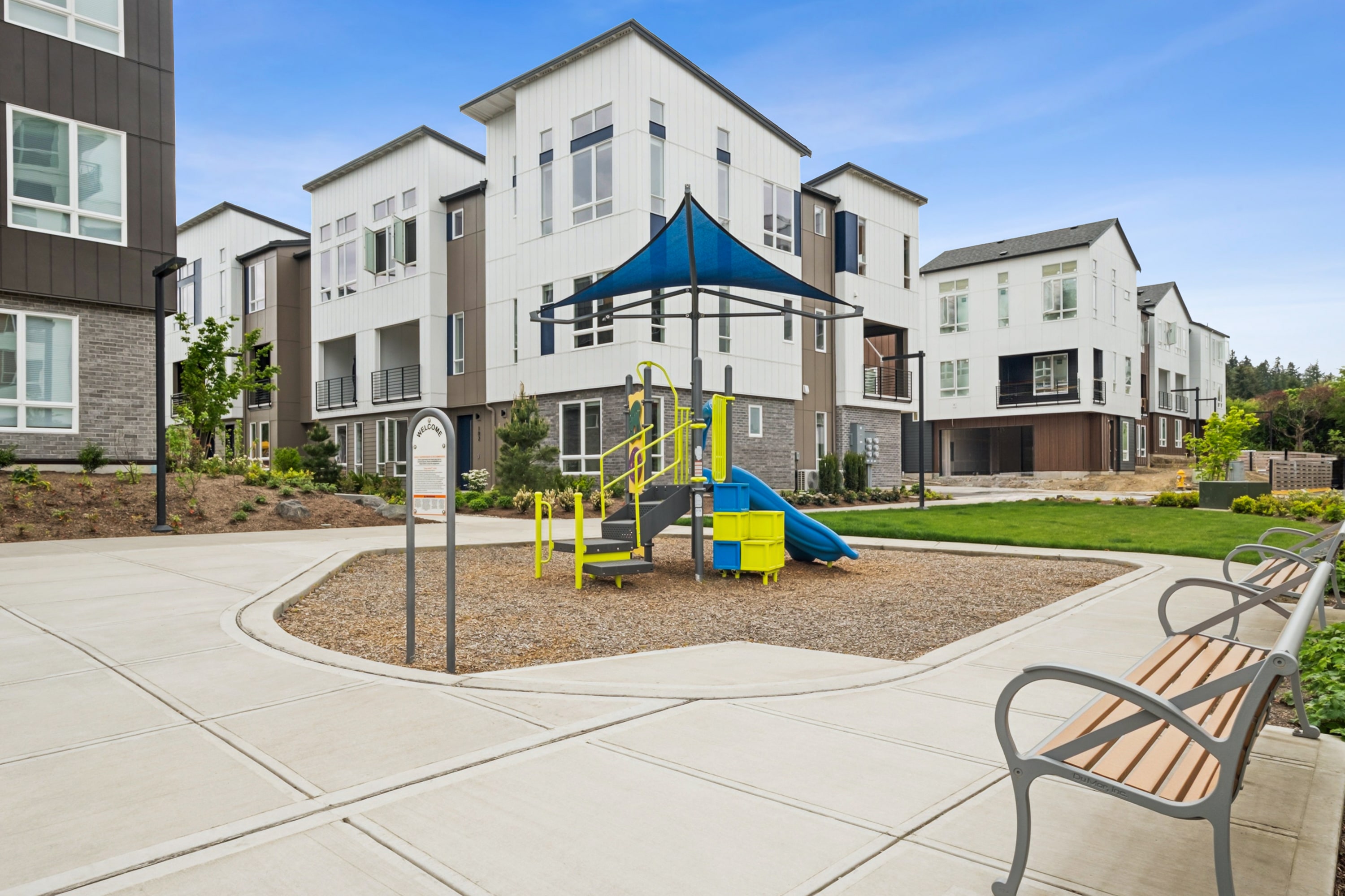 A modern residential complex with a playground in the foreground, surrounded by multi-story buildings with a mix of architectural styles and colors.