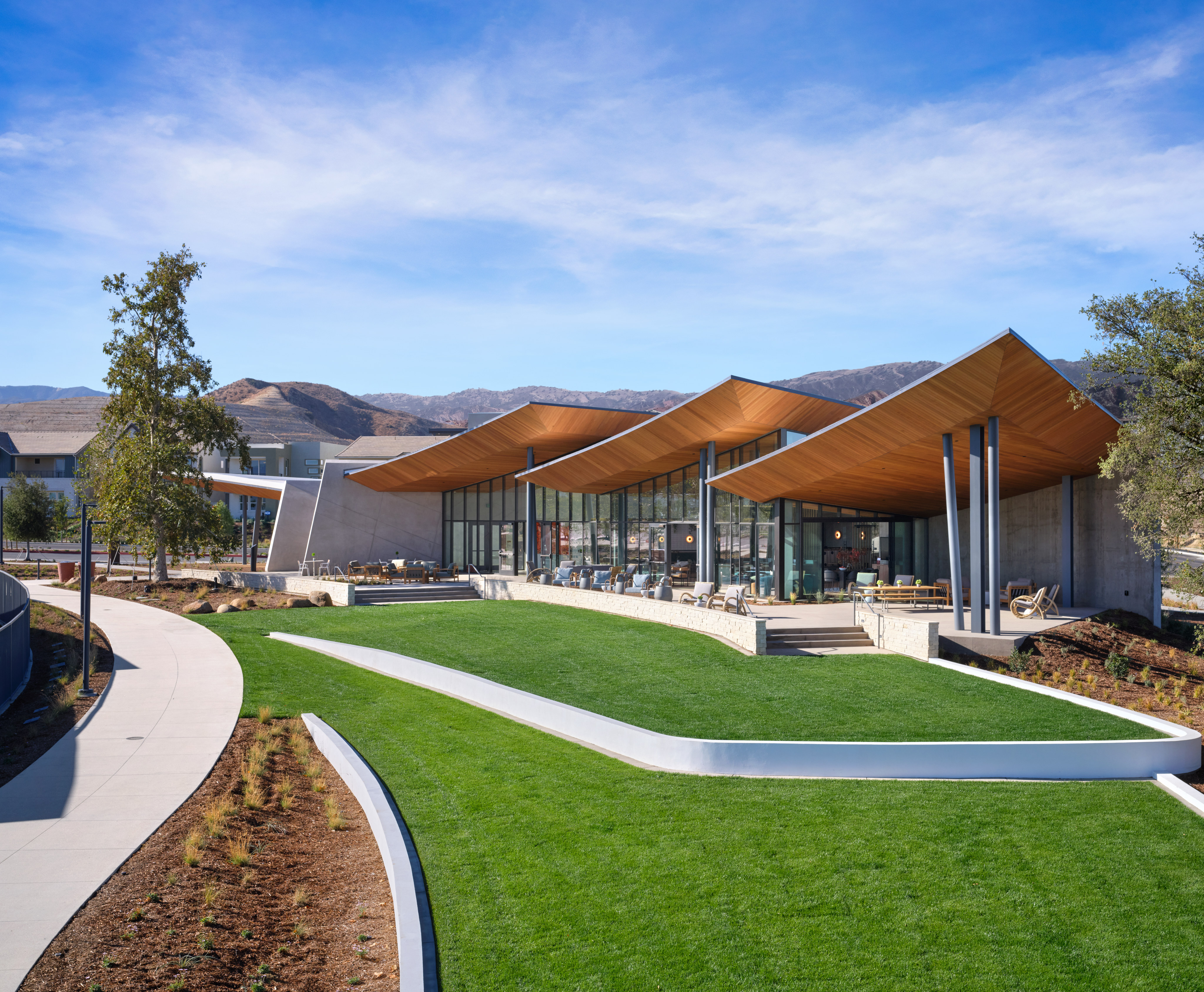 A modern, architecturally-designed building with a curved, orange-colored roof stands in the foreground, surrounded by a well-manicured lawn and landscaping. In the background, a mountainous landscape can be seen under a clear, blue sky.