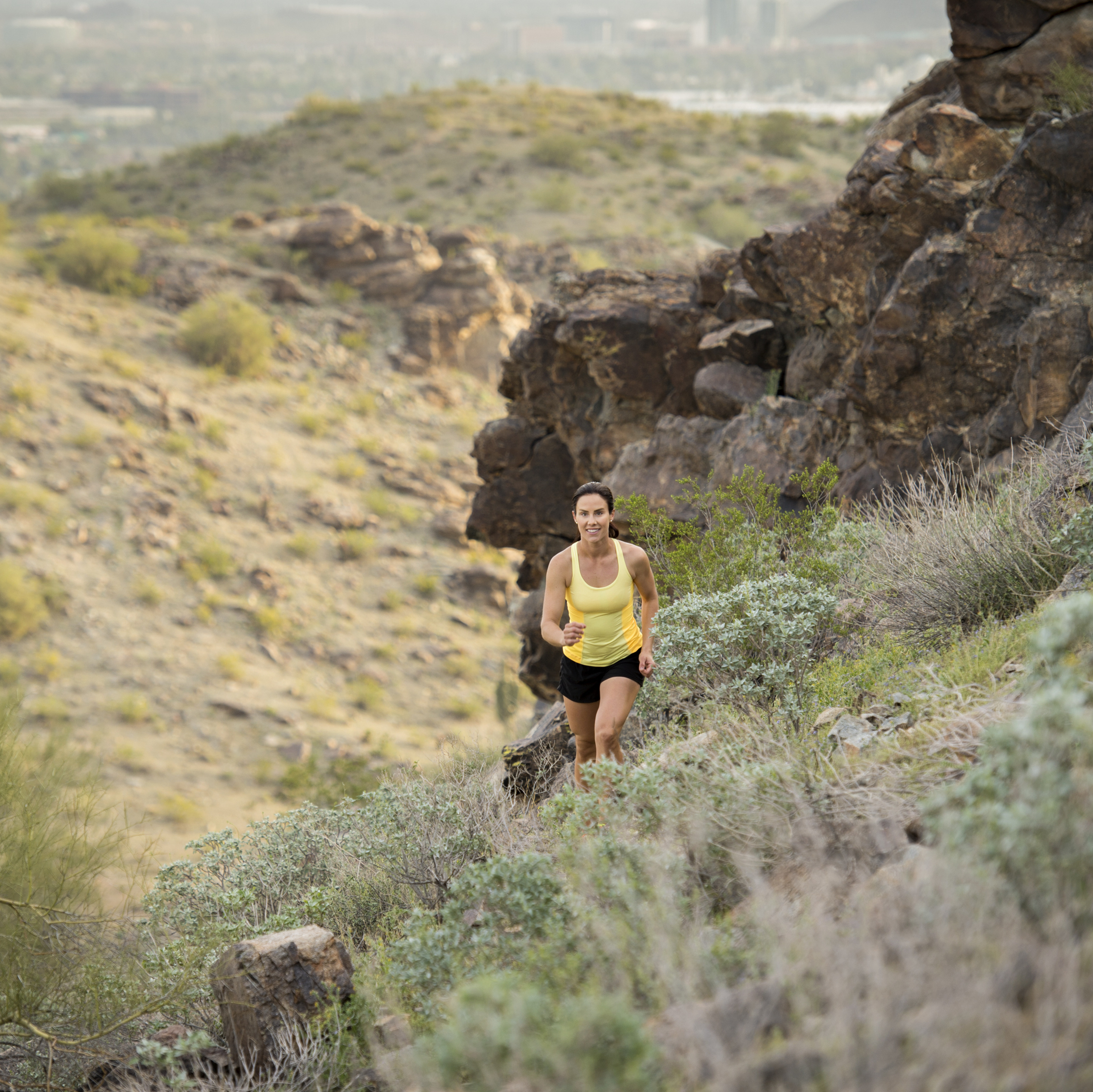 A person in athletic clothing is running on a rocky trail in a mountainous landscape with a city skyline visible in the distance.