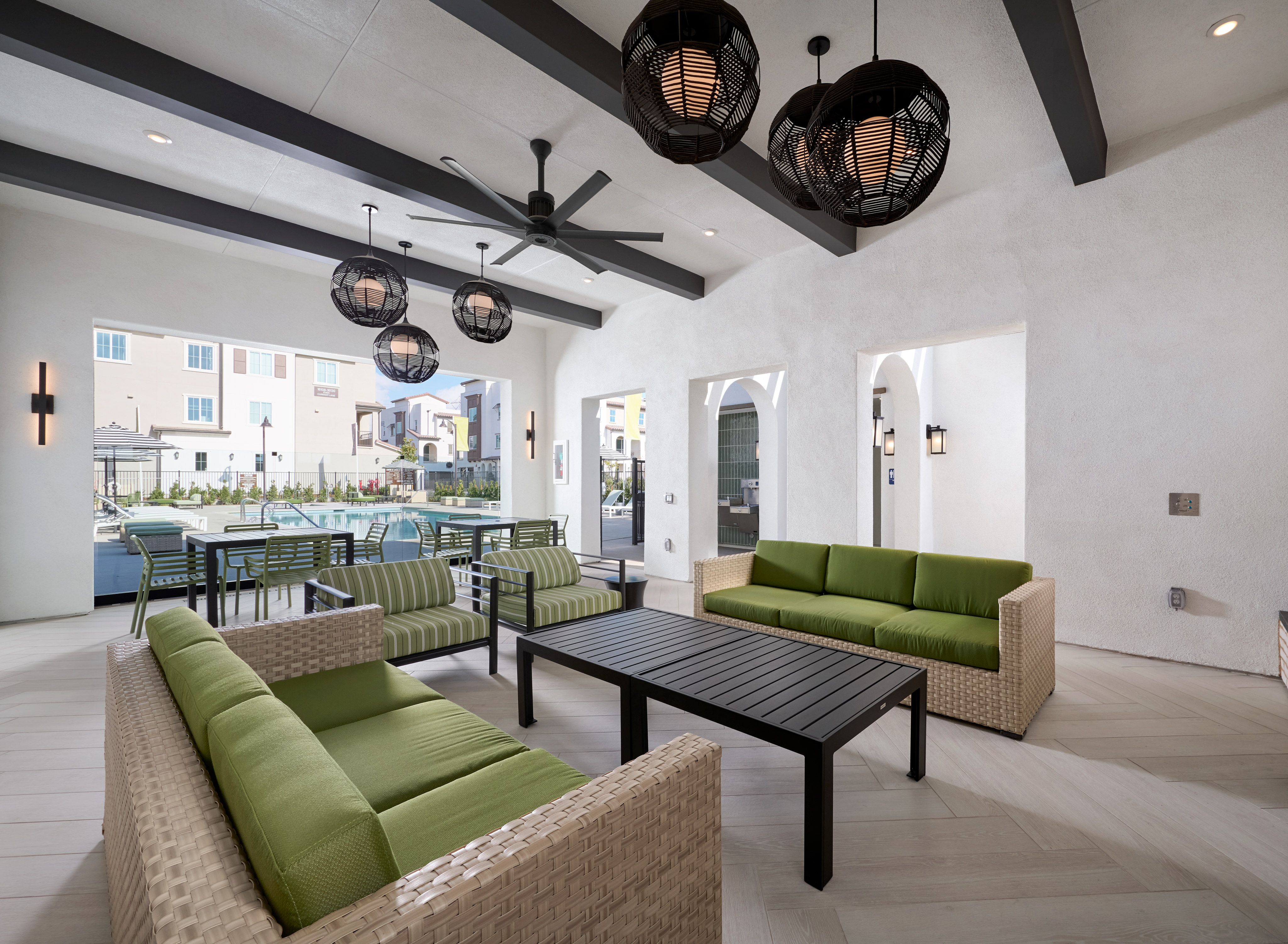 A modern and stylish living room with comfortable green sofas, a black coffee table, and hanging light fixtures, set against a backdrop of a kitchen area with a bar counter and appliances.