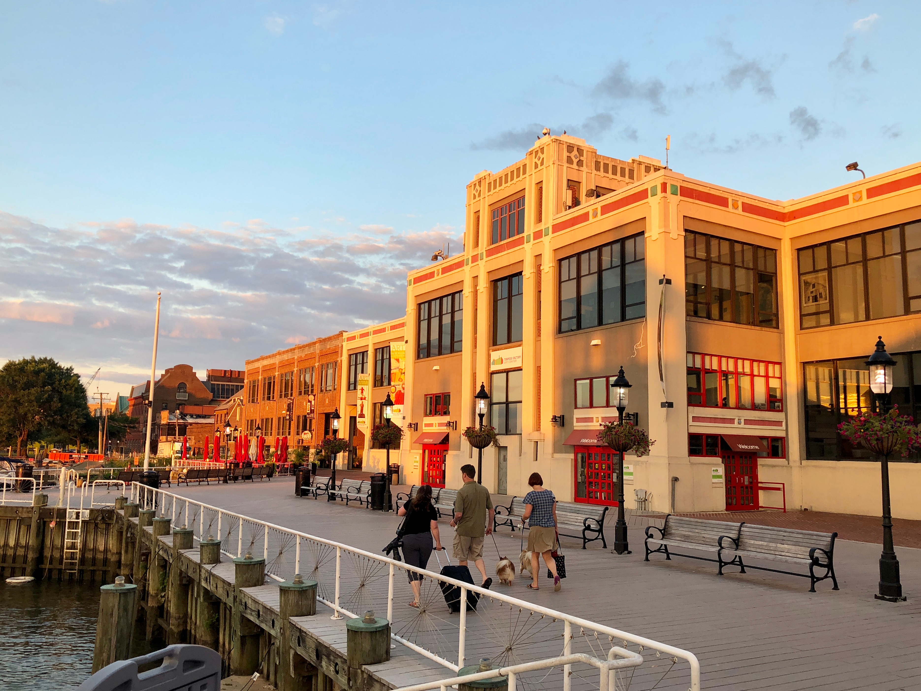 A vibrant waterfront scene with a colorful, ornate building in the foreground and people walking along the boardwalk, set against a backdrop of a cloudy sky.