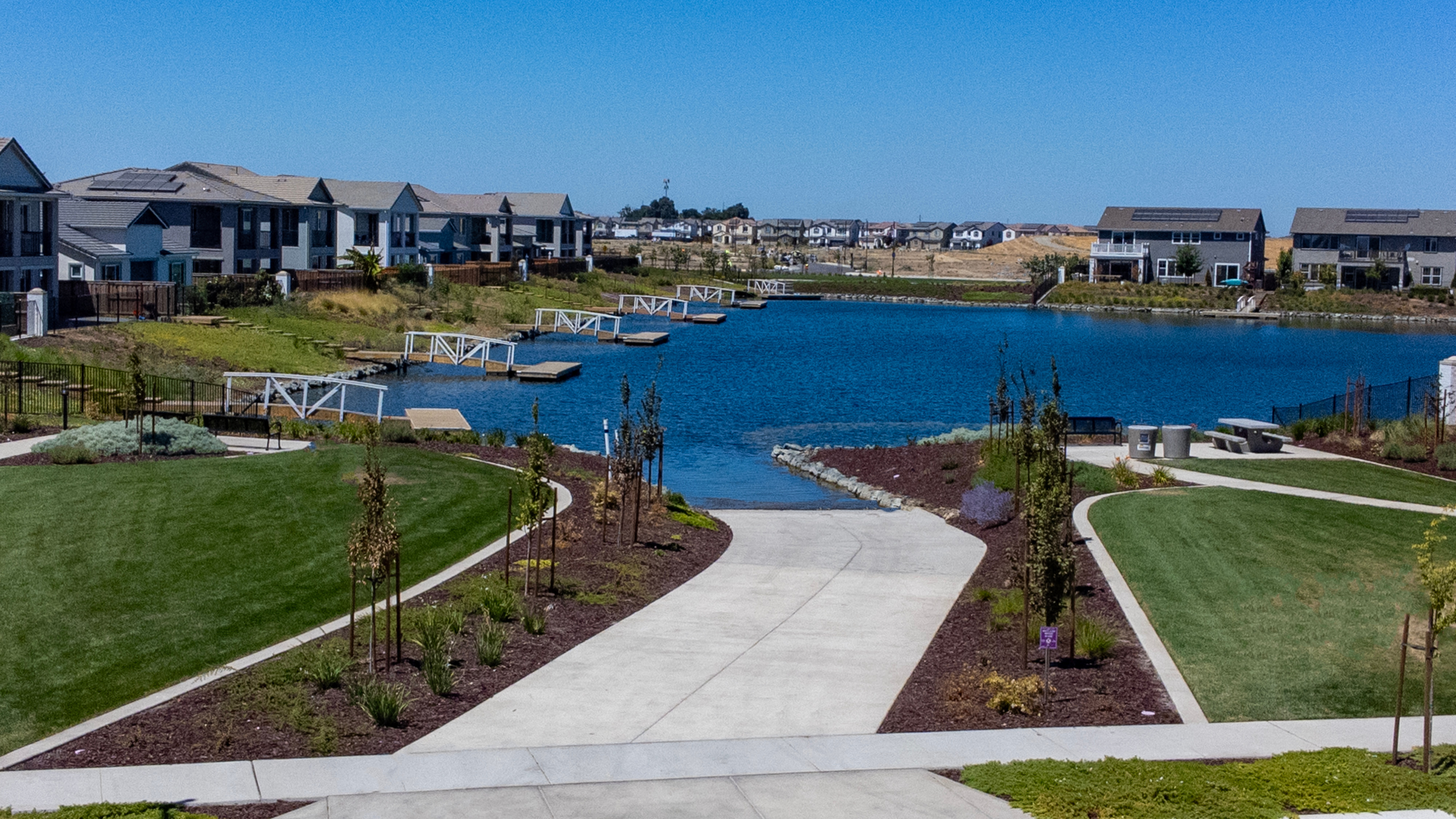 A well-manicured walkway leads through a lush, landscaped community with a picturesque lake and surrounding residential buildings in the background.