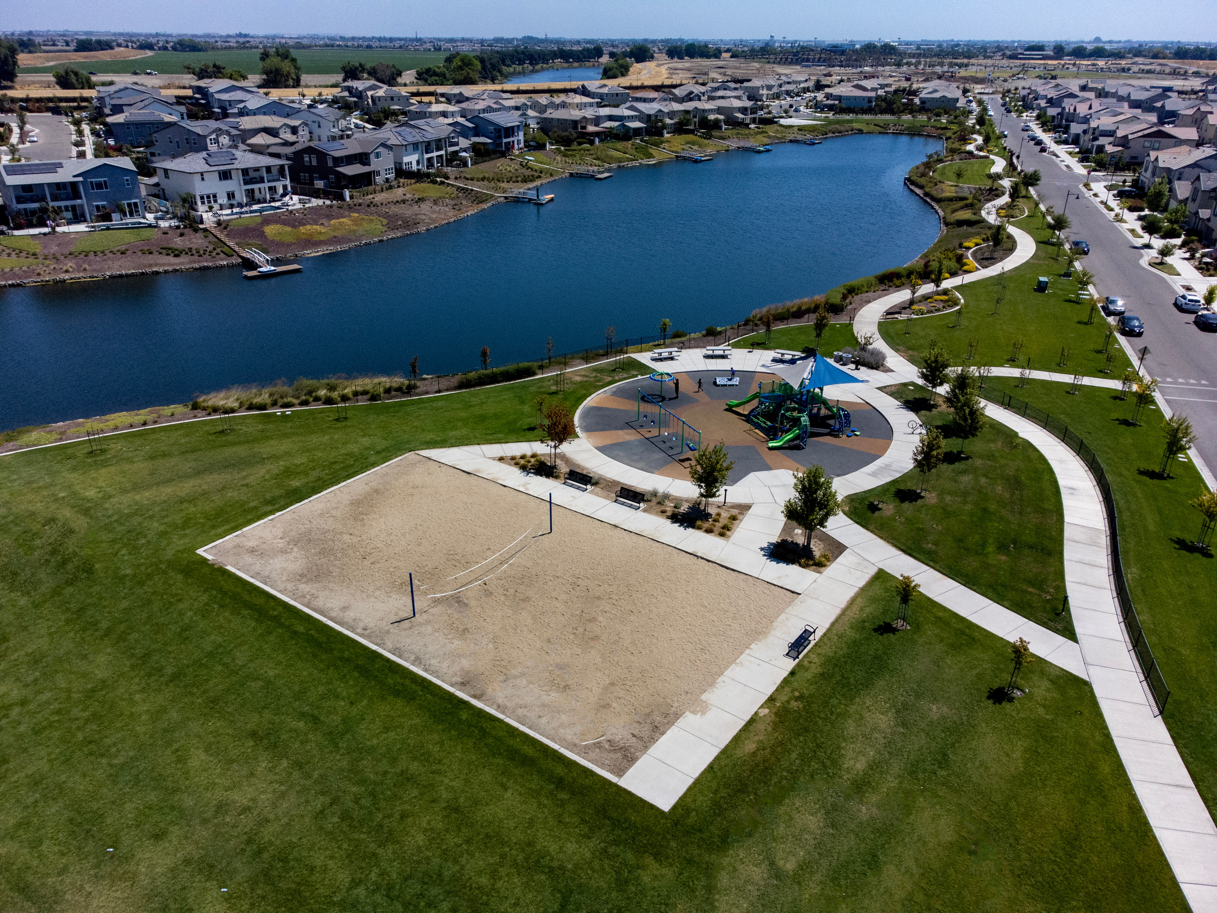 A residential community with a central lake, surrounded by well-manicured lawns, walking paths, and a decorative fountain in the foreground.