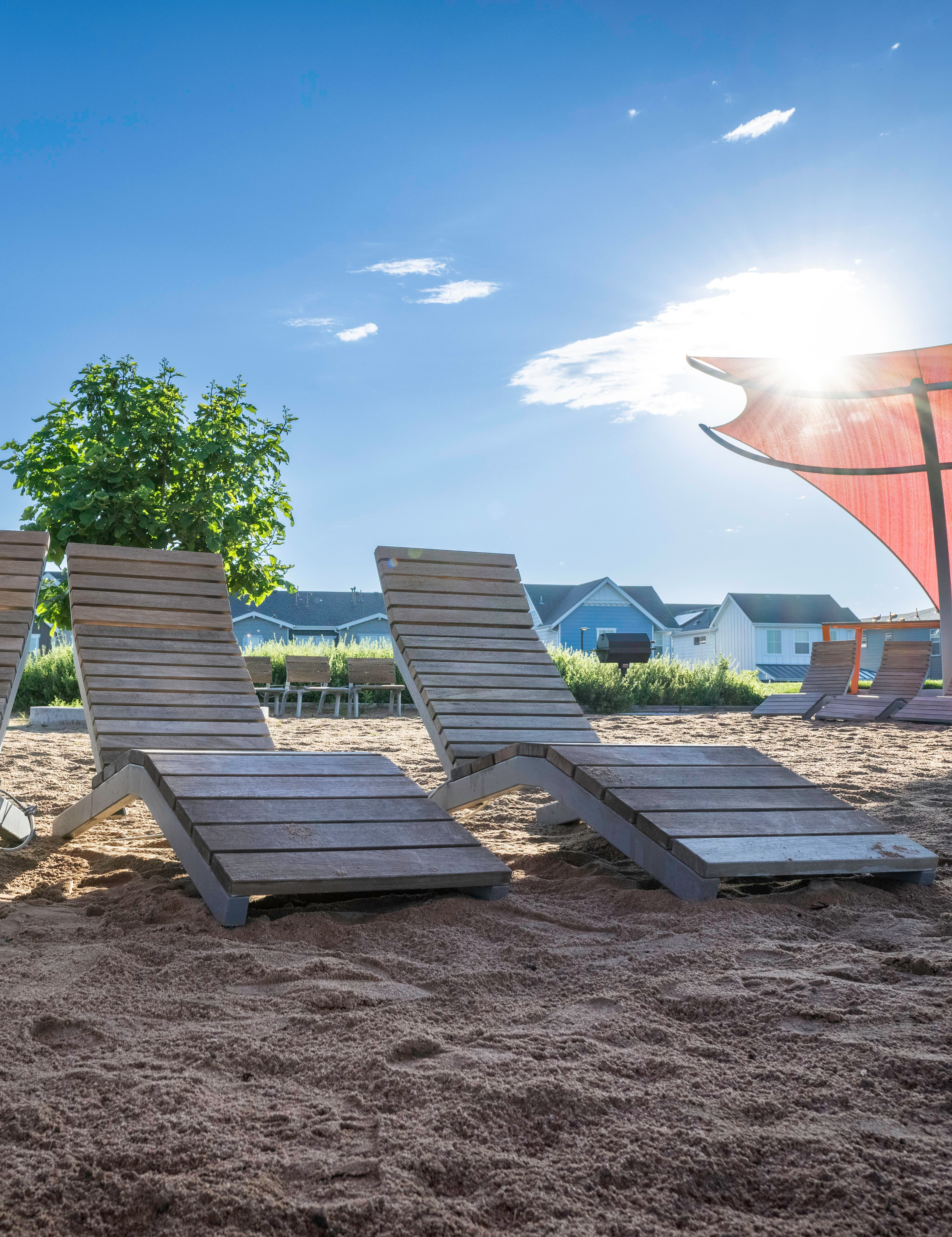 Wooden lounge chairs arranged on a sandy beach, with a vibrant red umbrella providing shade and a lush green tree in the background against a clear blue sky.
