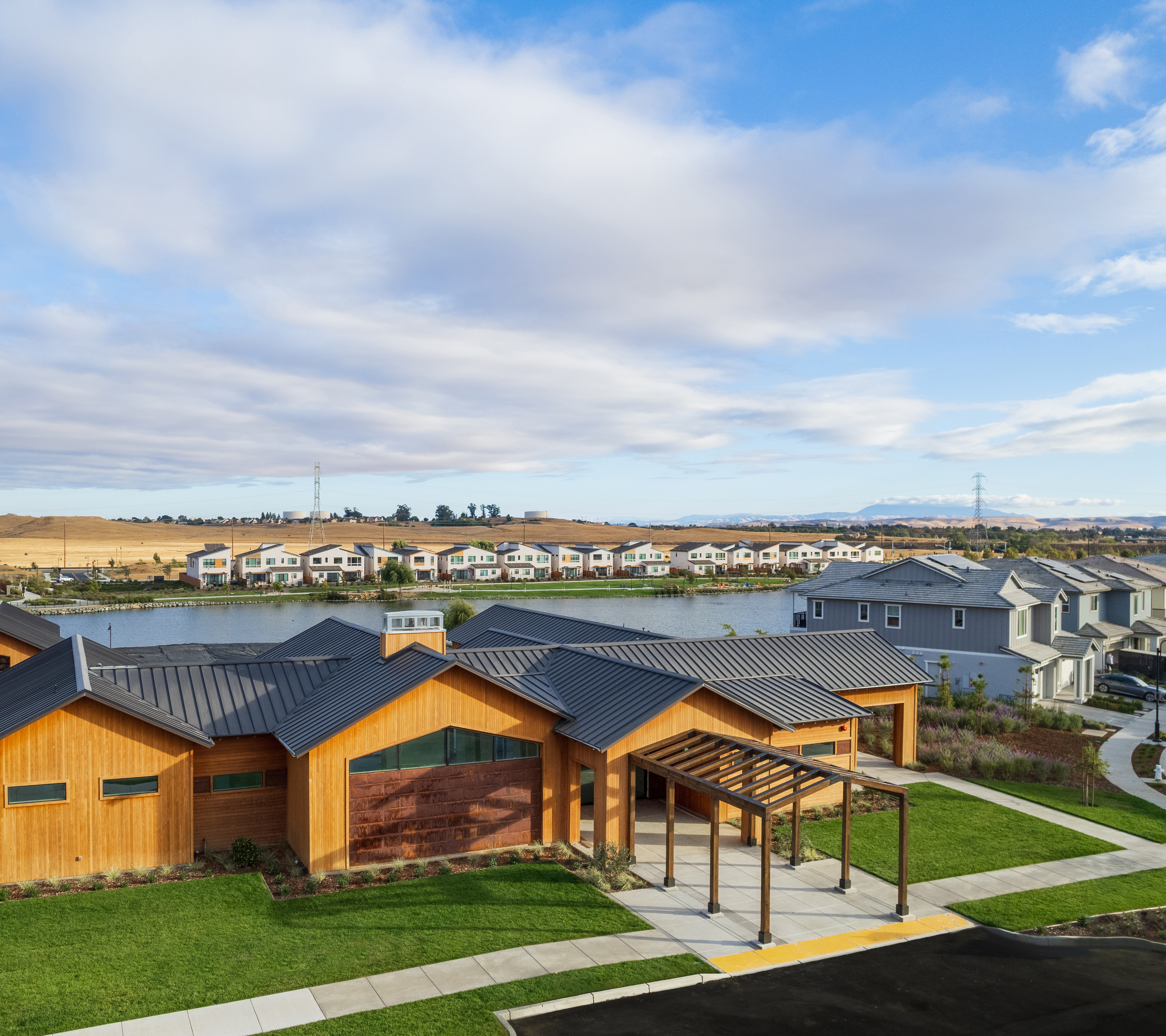A residential neighborhood with colorful wooden houses surrounded by lush green lawns, set against a backdrop of a cloudy blue sky.