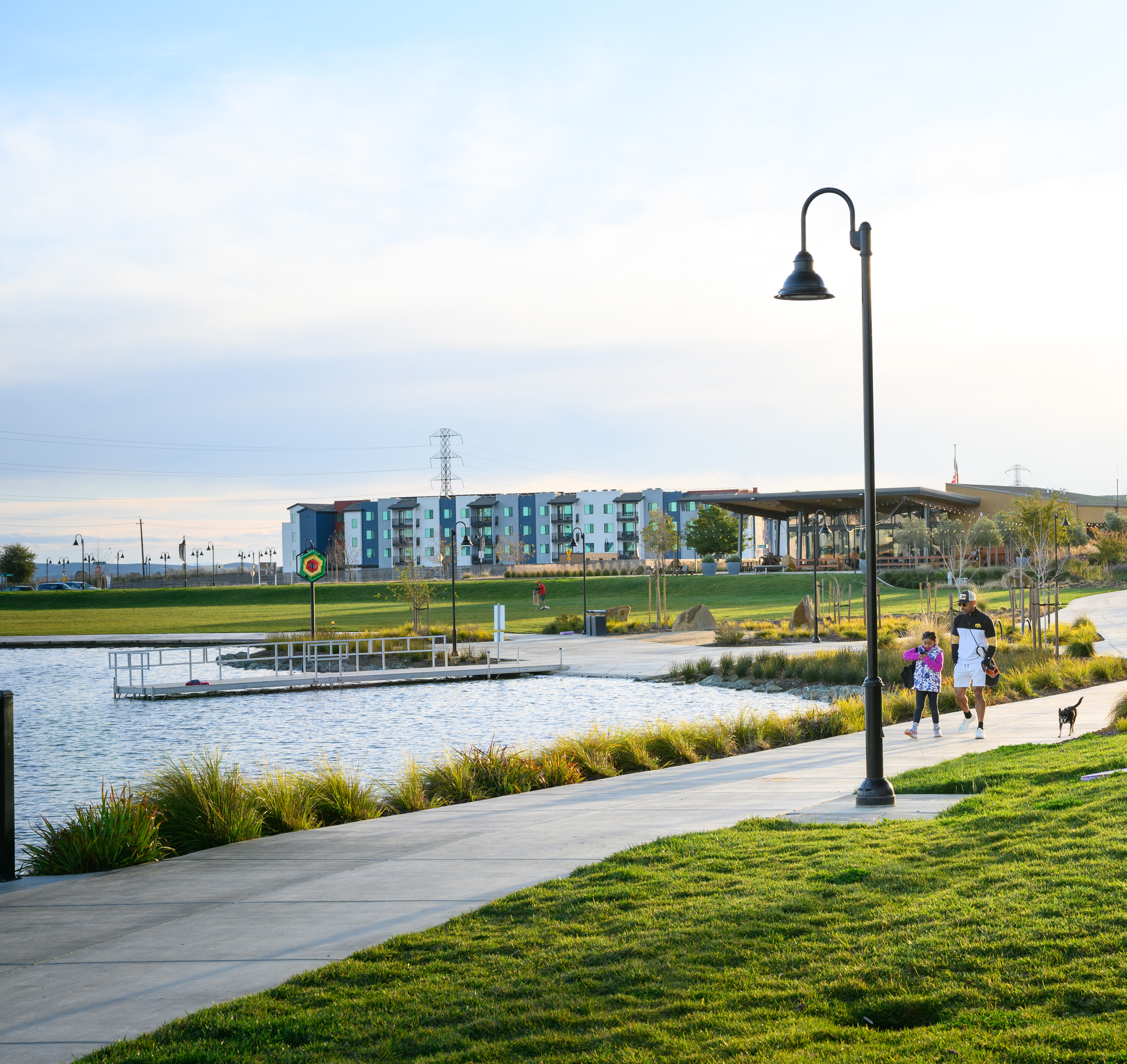 A scenic outdoor public space with a paved walkway, lush green grass, and a decorative lamp post, with a body of water and colorful buildings visible in the background.