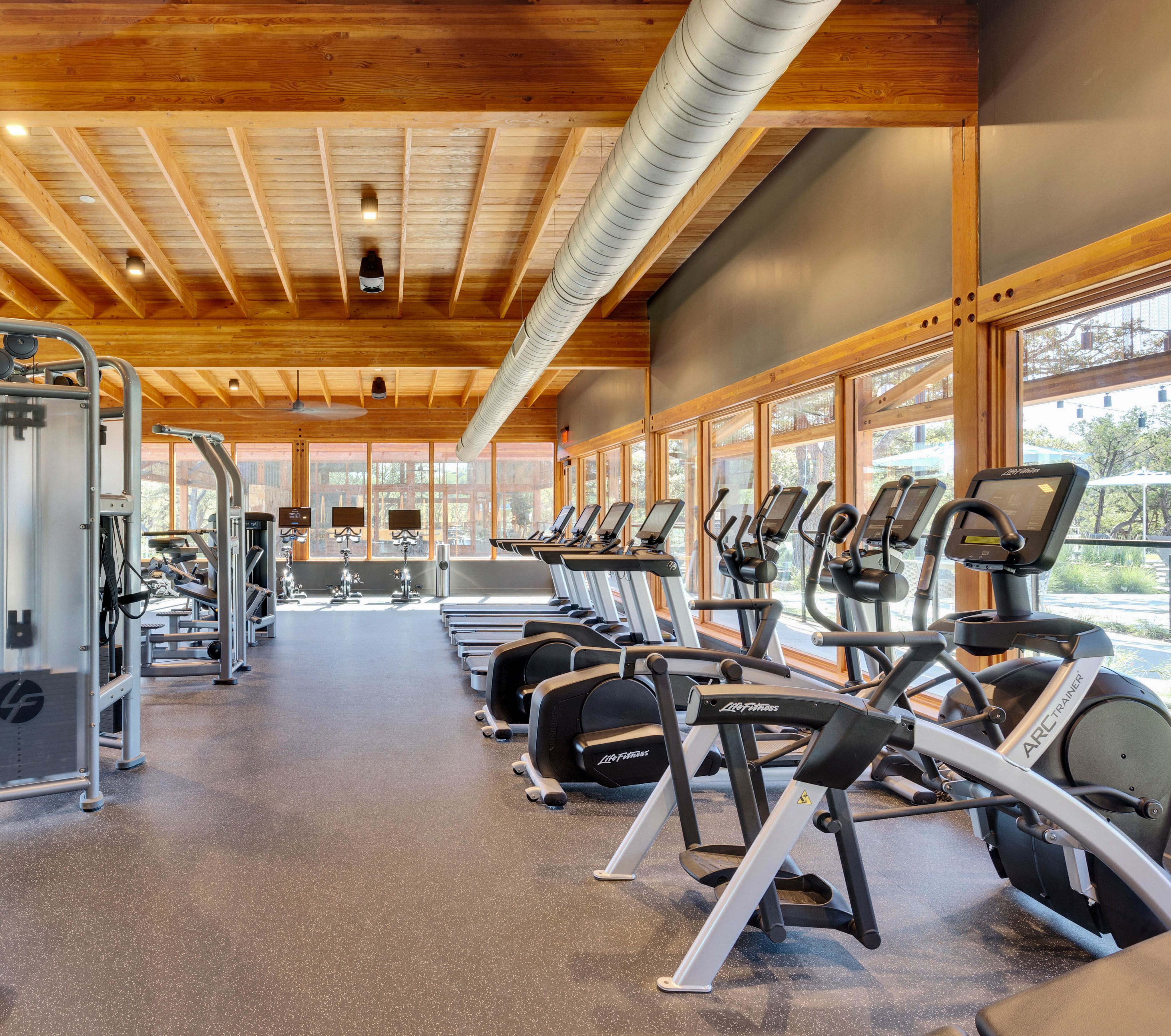 A well-equipped gym with various exercise equipment, including treadmills, elliptical machines, and weight training machines, set against a backdrop of wooden beams and large windows providing natural light.