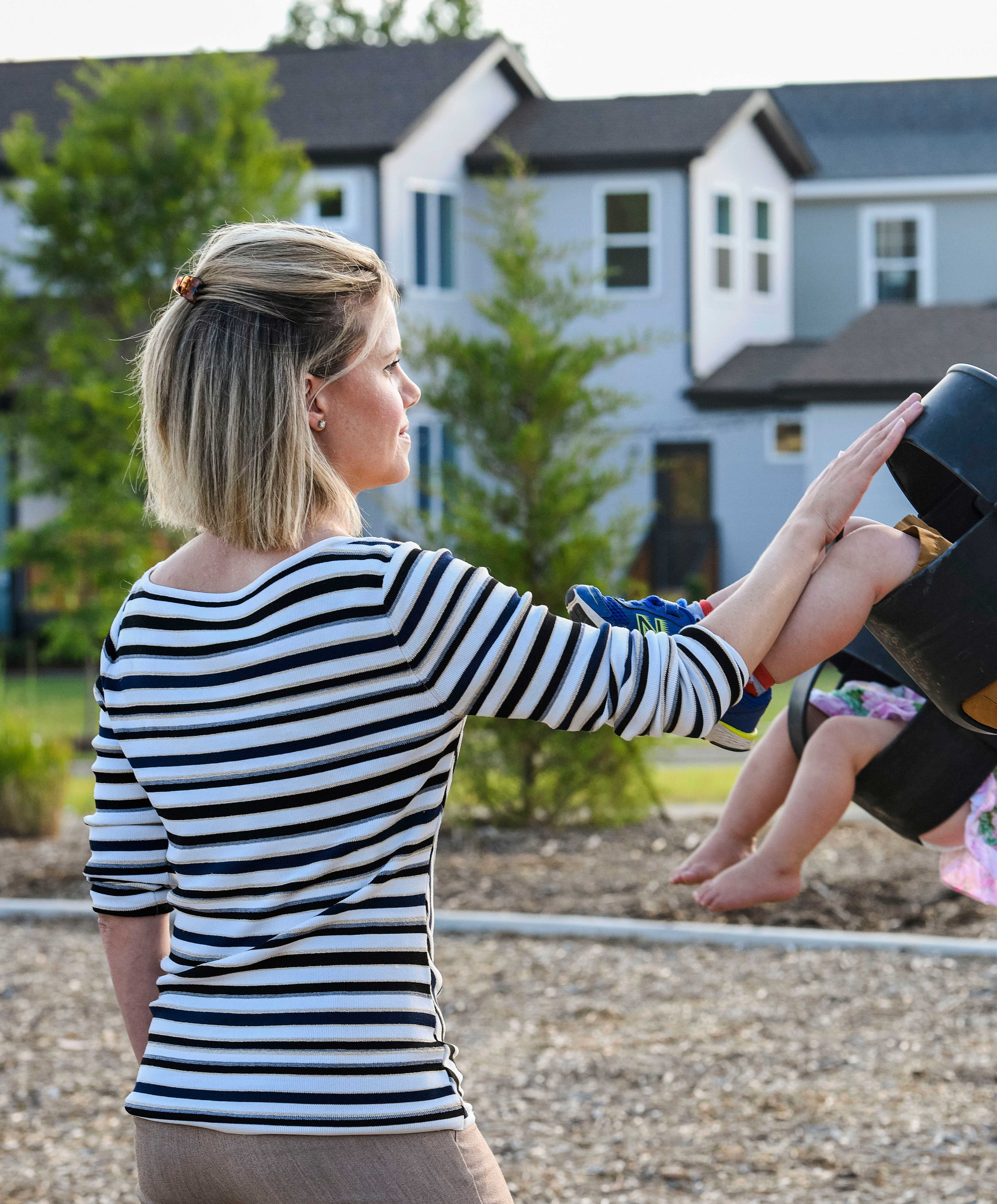 A woman in a striped shirt is pushing a young child on a swing in a playground surrounded by residential buildings.