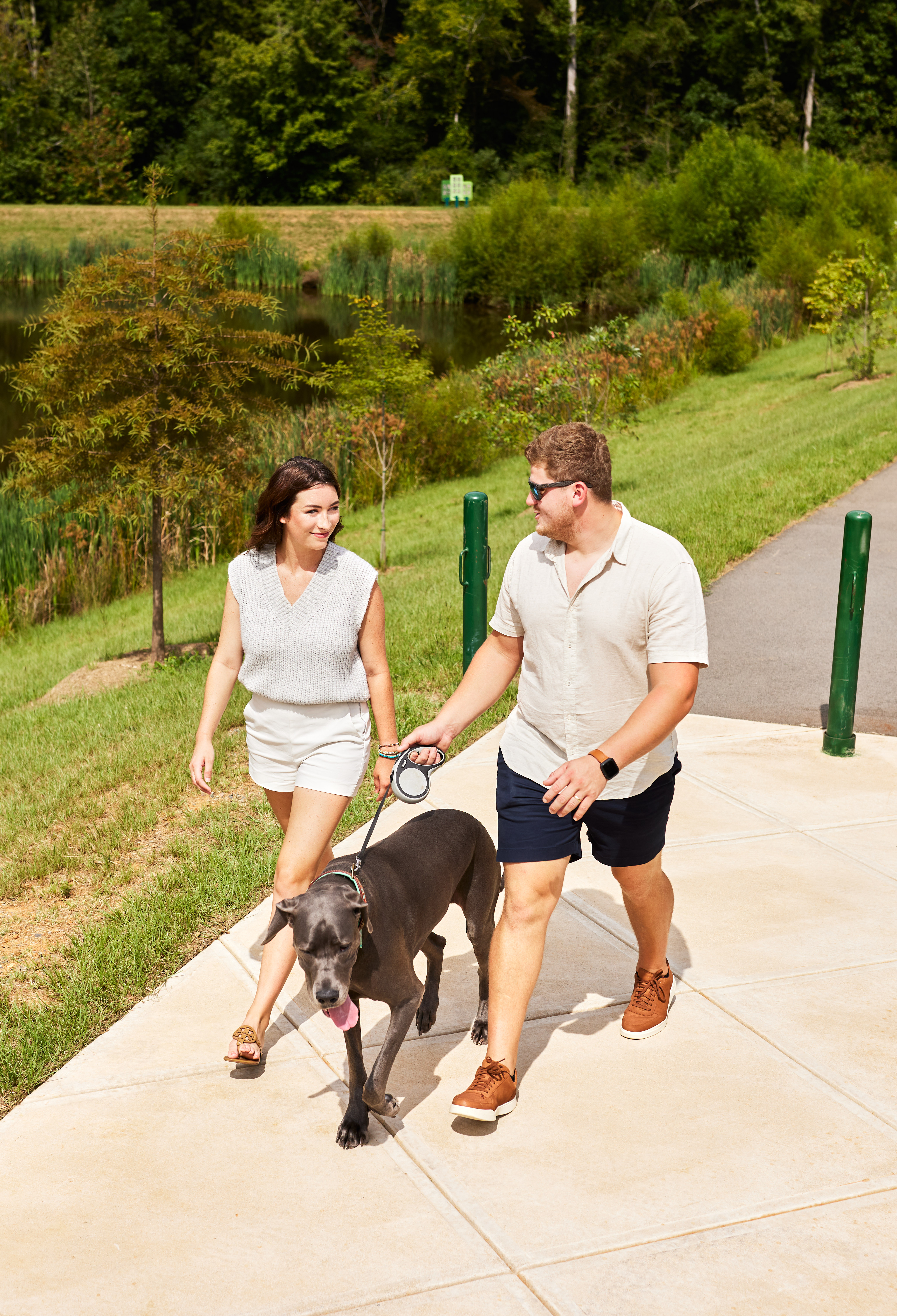 A couple walks their dog along a paved path surrounded by lush greenery, with a pond visible in the background.