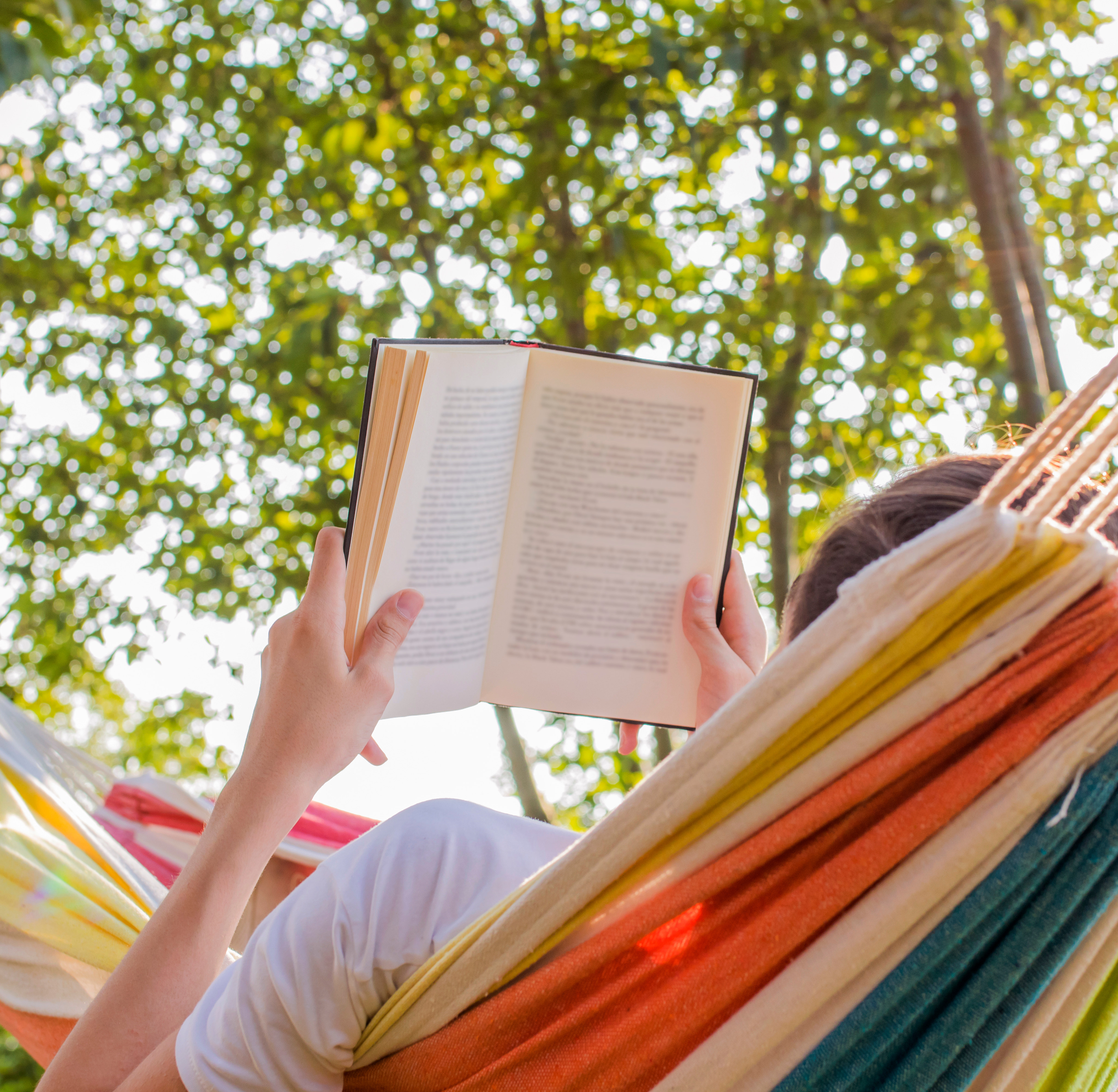 A person is relaxing in a colorful hammock, reading a book amidst the lush greenery of the surrounding trees.