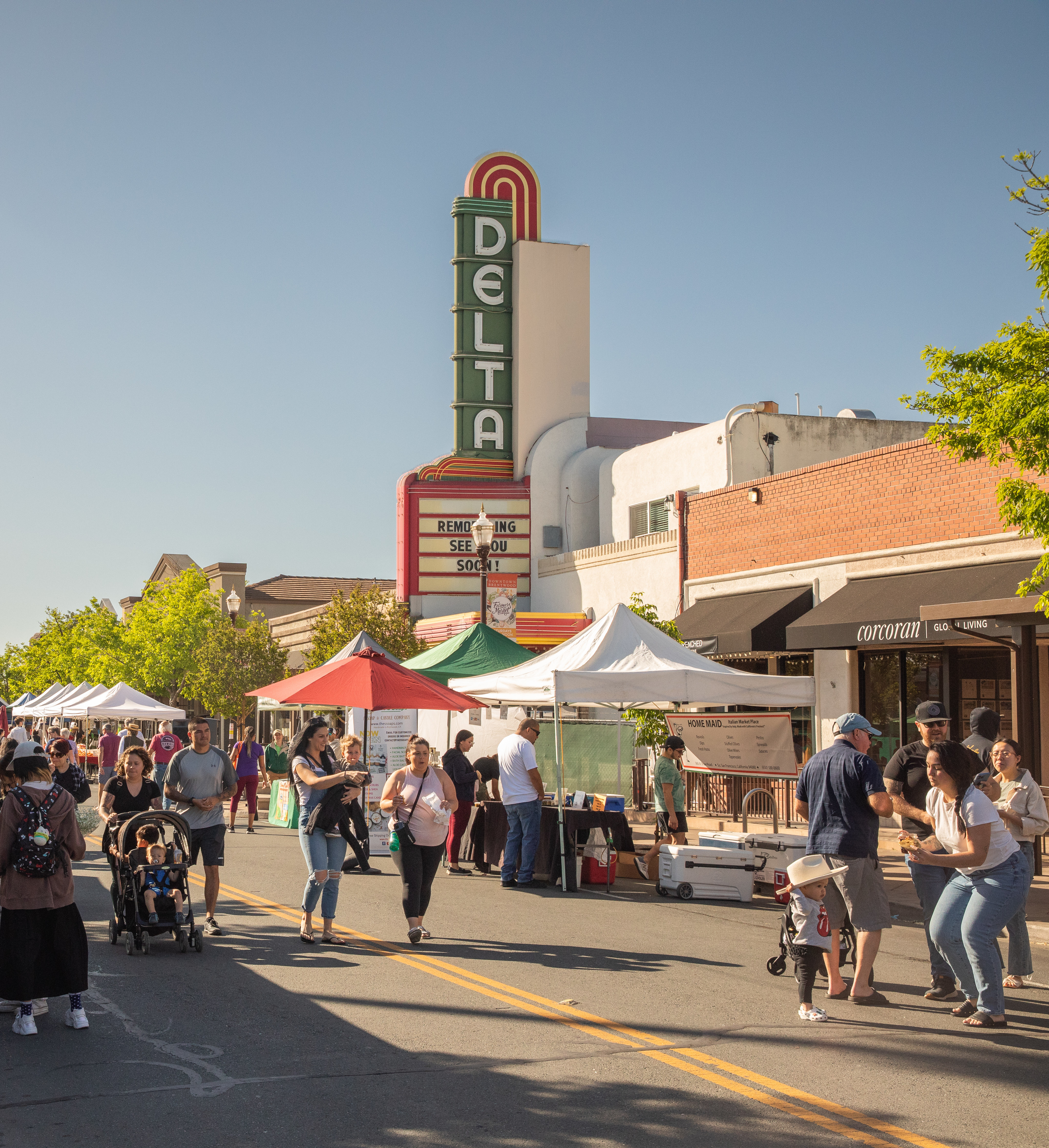 A bustling outdoor market with various vendors and tents, set against the backdrop of a historic Delta theater building with its iconic neon sign.