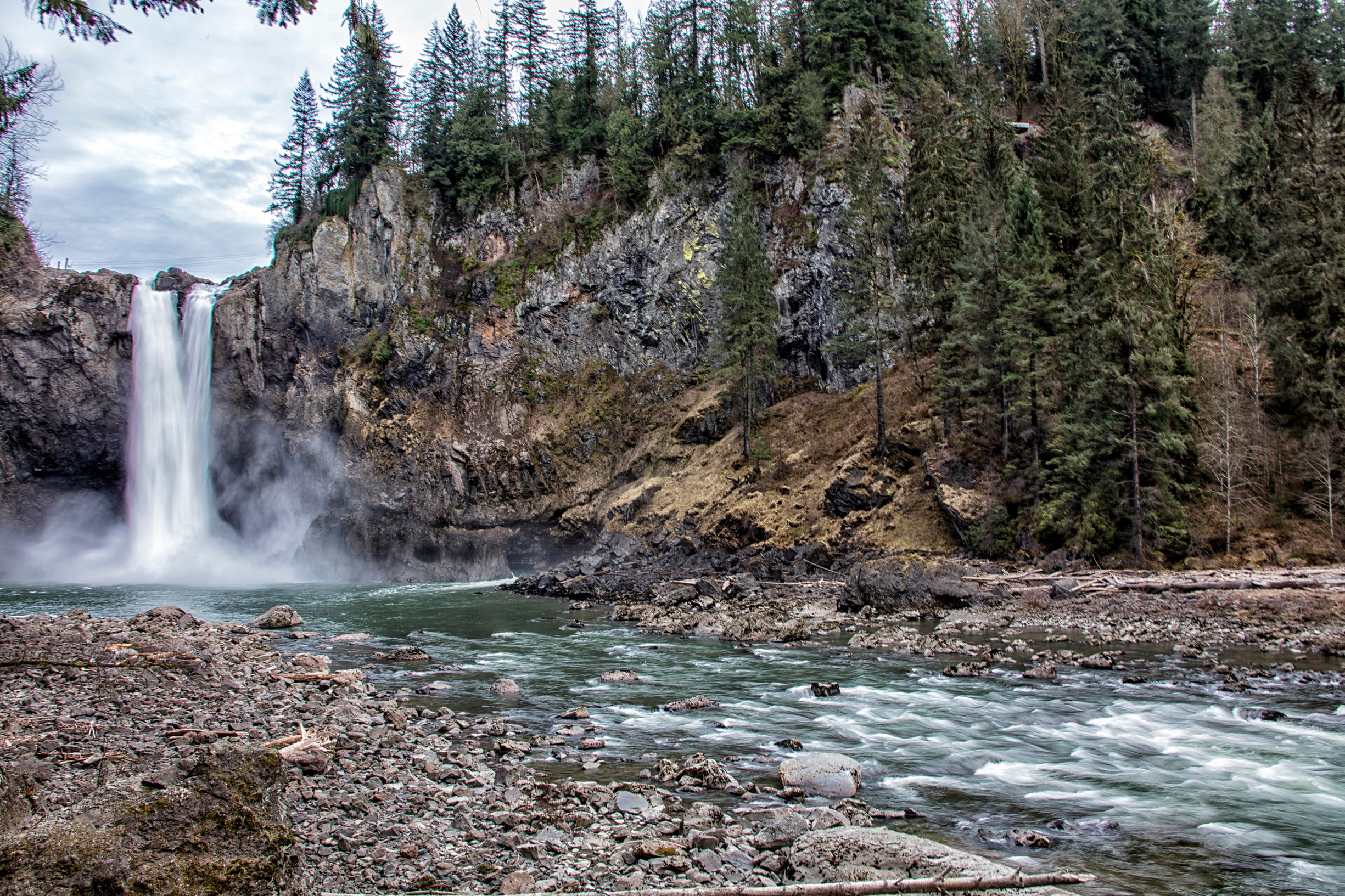 A majestic waterfall cascades down a rugged, forested cliff, with a swift-flowing river in the foreground and a lush, evergreen landscape in the background.