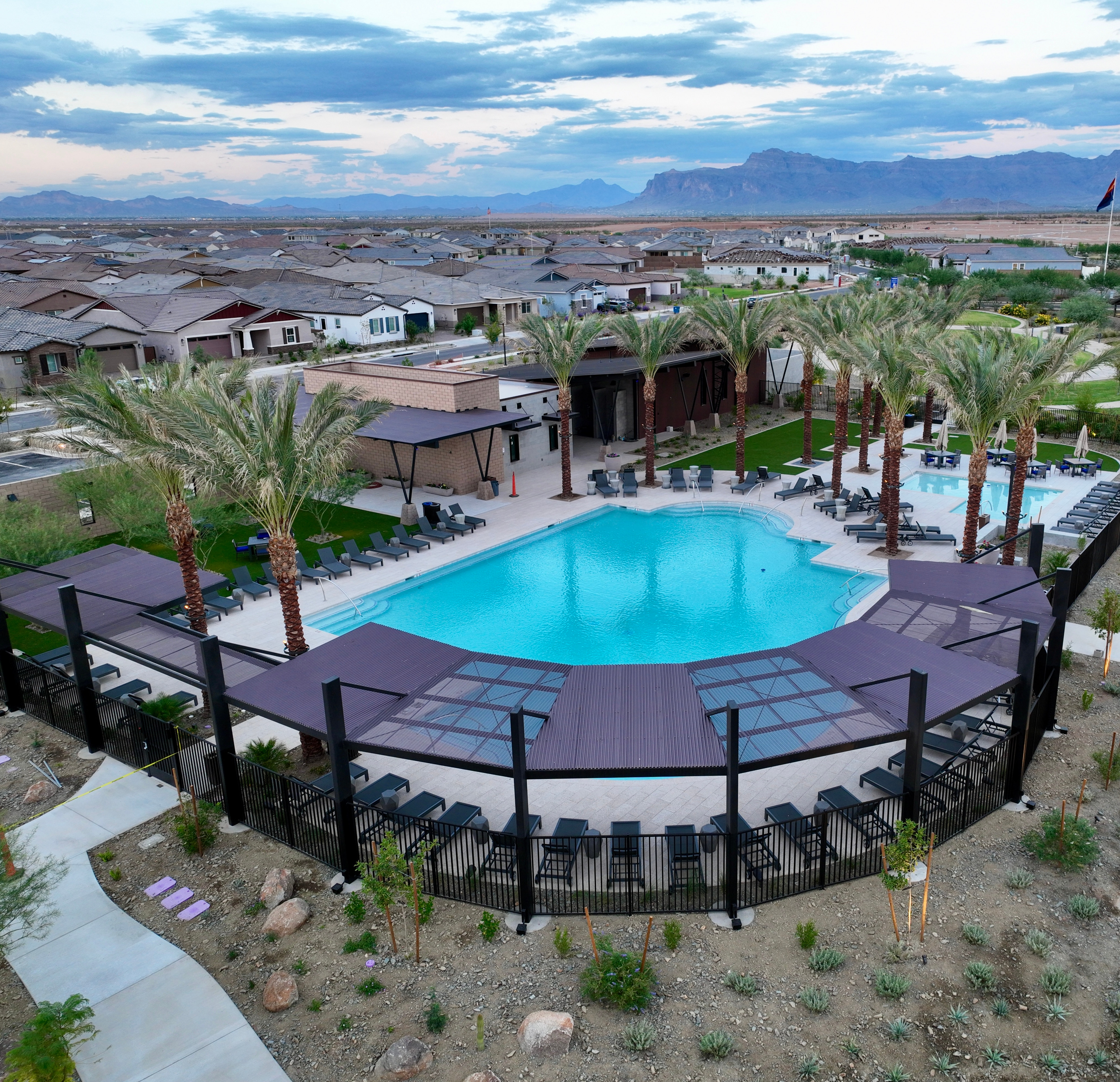 A large outdoor swimming pool surrounded by palm trees and buildings, with a mountainous landscape visible in the background.