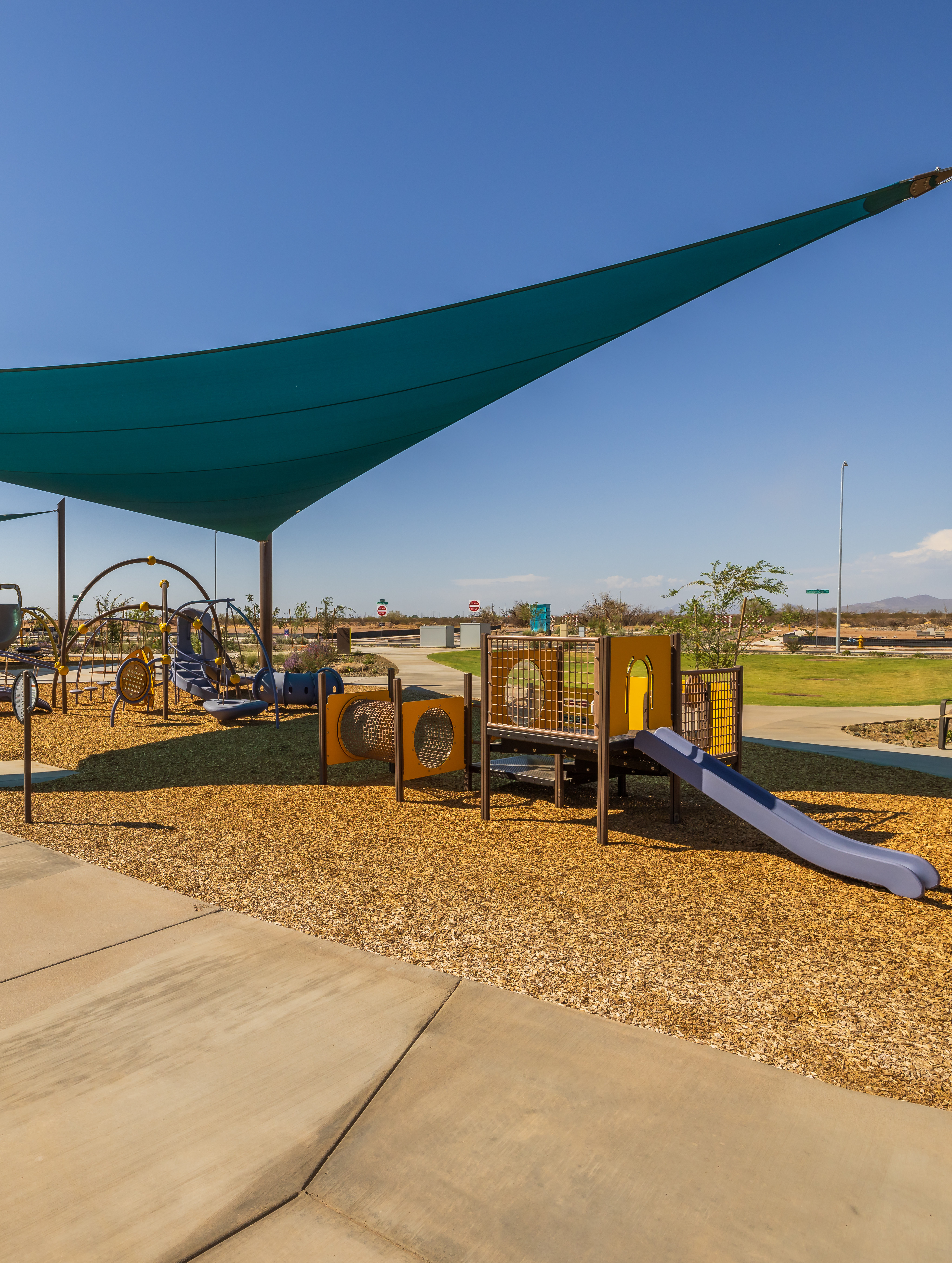 A shaded playground area with various play equipment, surrounded by a grassy landscape and a clear blue sky.