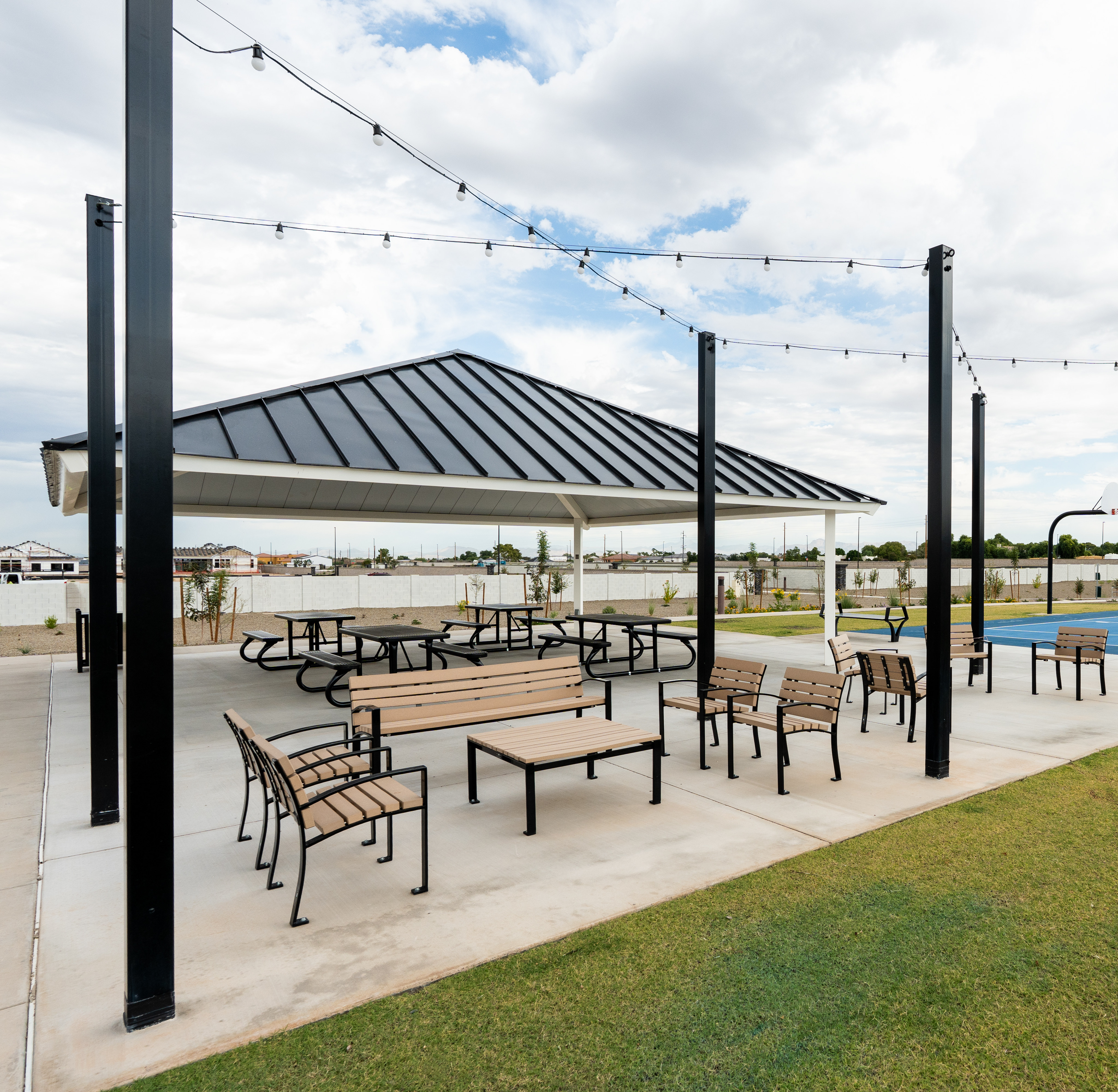 A covered outdoor seating area with tables and chairs, surrounded by a grassy lawn and a swimming pool in the background, with string lights overhead and a cloudy sky above.