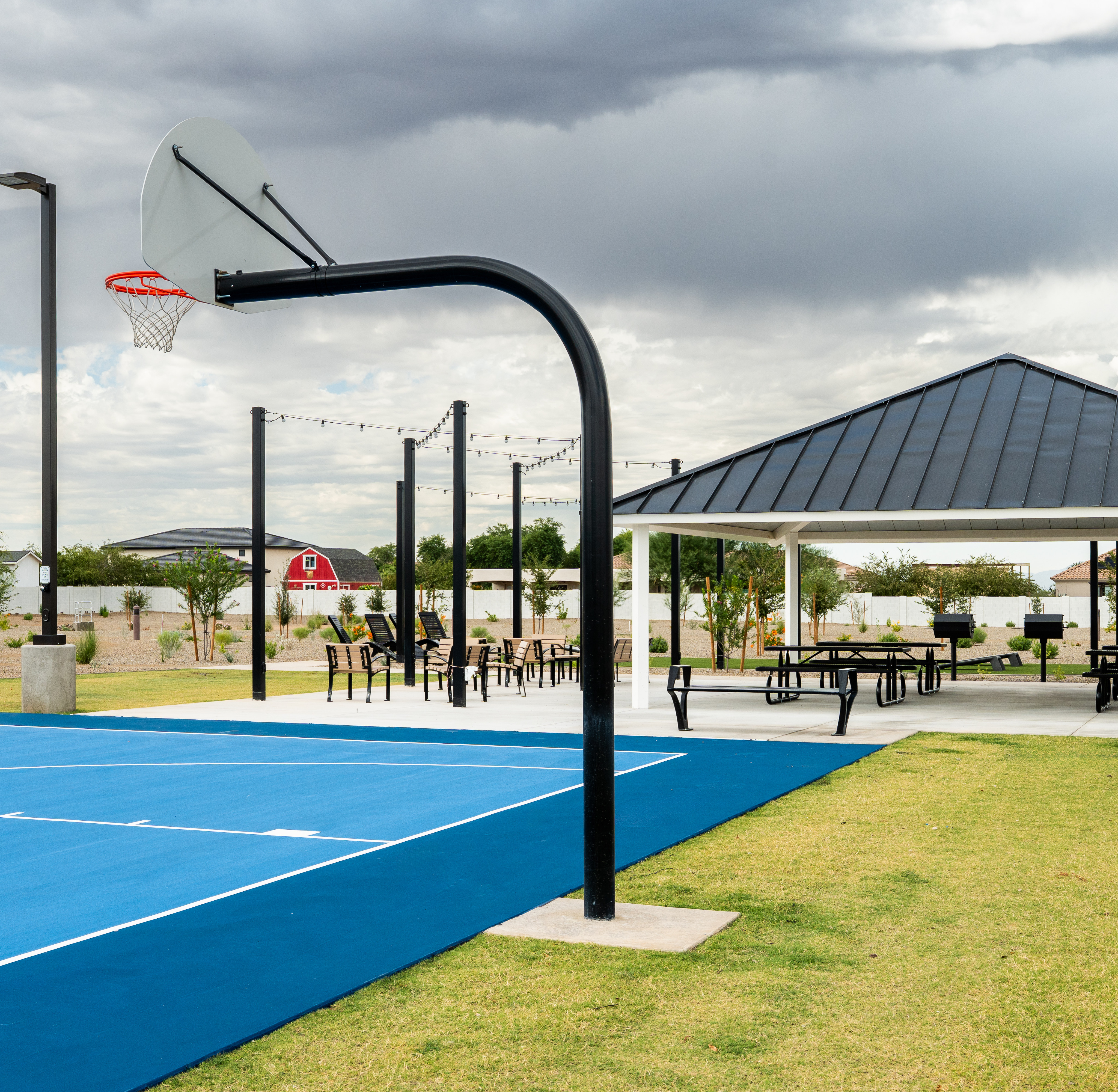 A basketball court with a hoop and net stands in the foreground, surrounded by a grassy area and a covered pavilion structure in the background against a cloudy sky.
