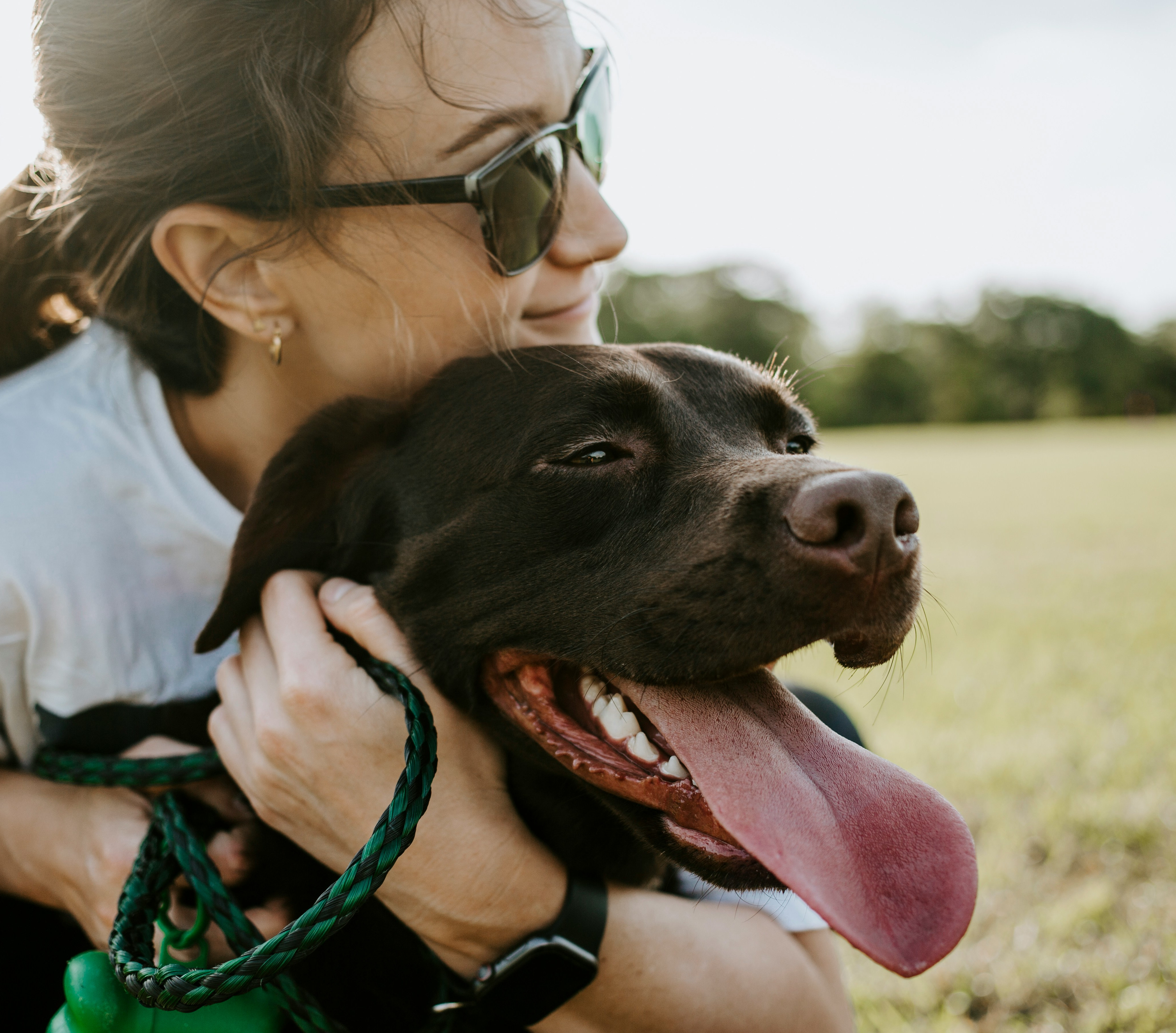 A woman in sunglasses is embracing a happy, black dog in a grassy field.