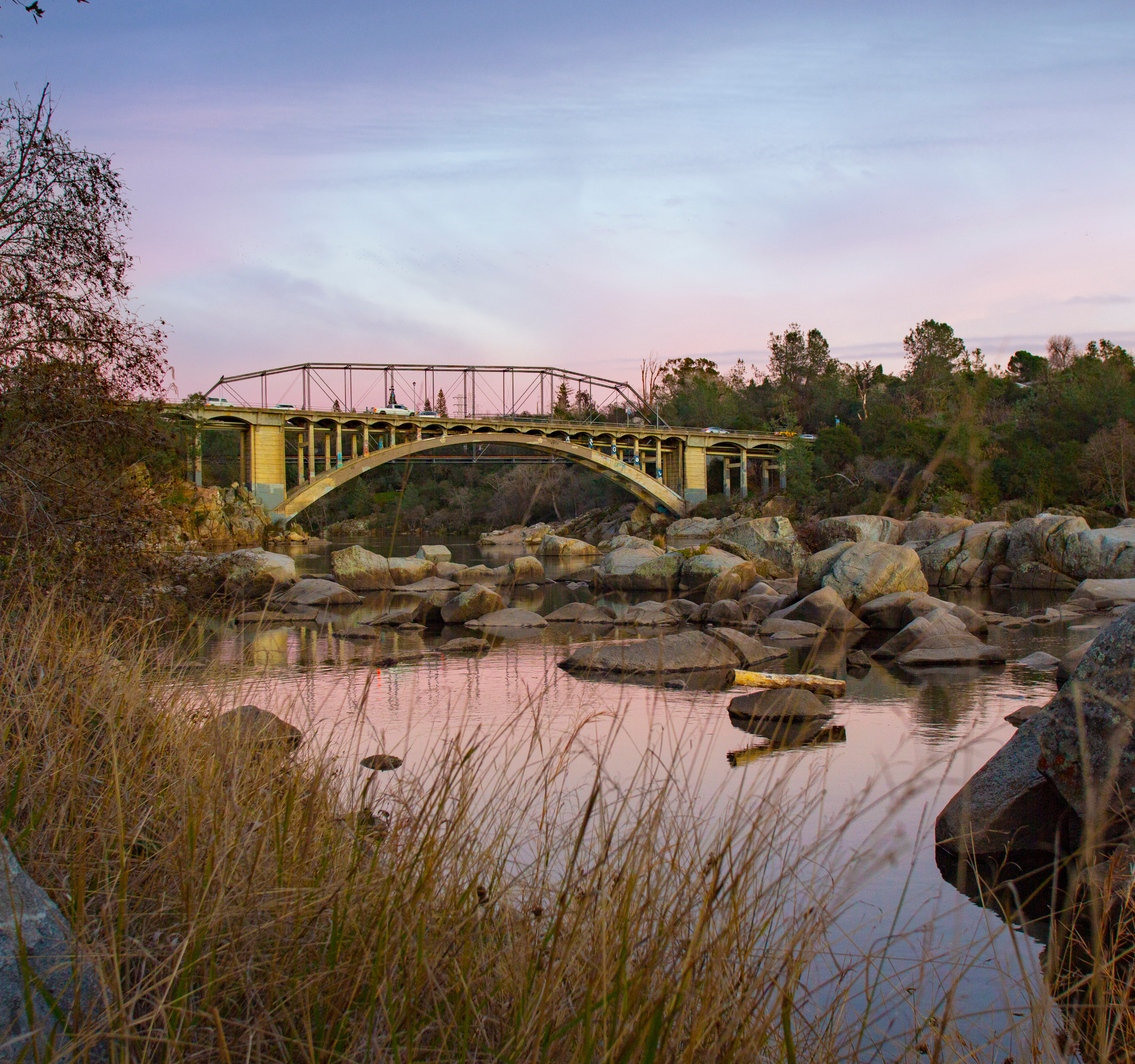 A tranquil pond reflects the arched bridge and surrounding trees and vegetation in a serene natural setting.