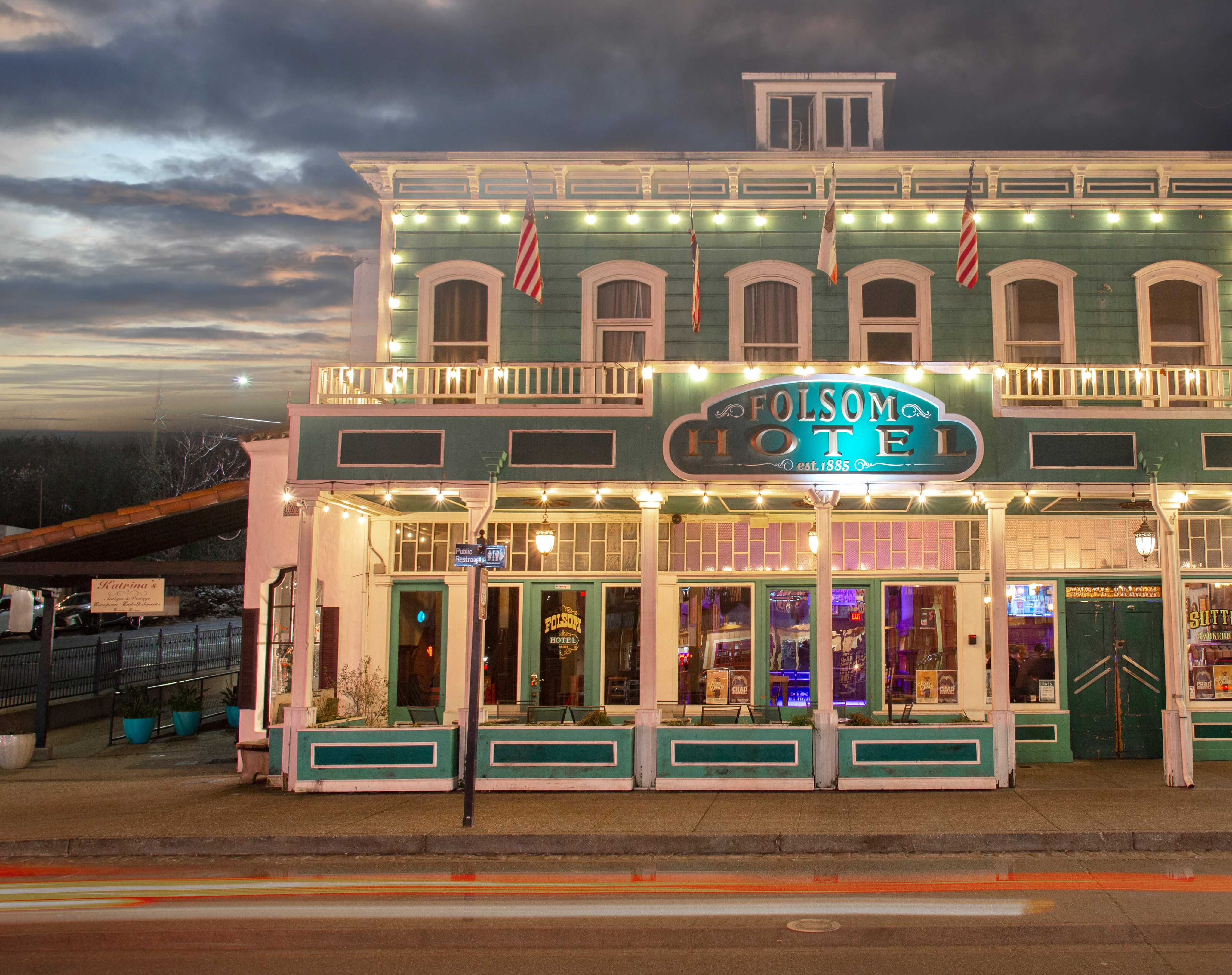 A colorfully lit and ornate hotel building stands in the foreground, with a cloudy sky and street lights visible in the background.