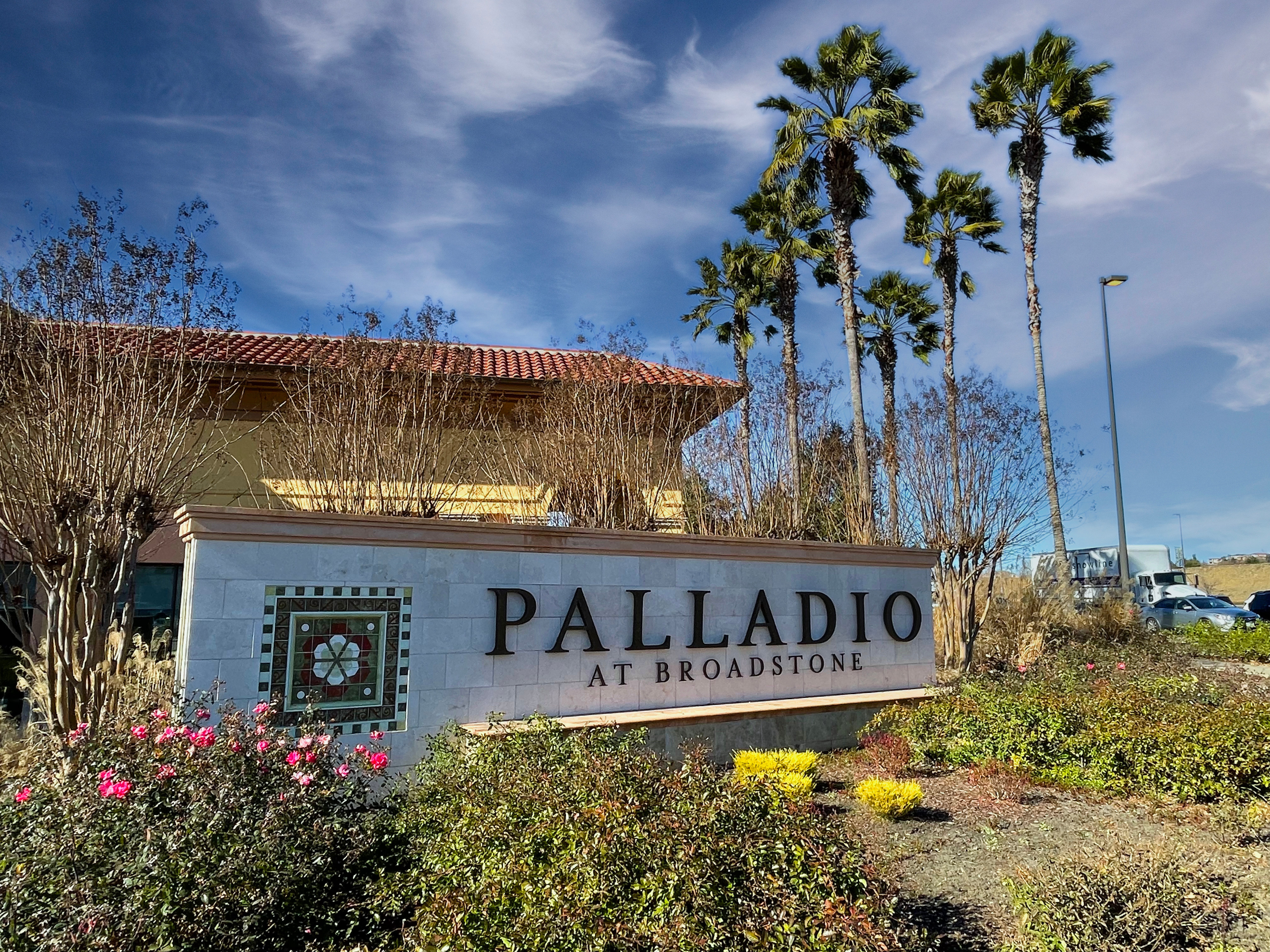 The image shows a sign for the Palladio at Broadstone, with palm trees and a blue sky in the background.