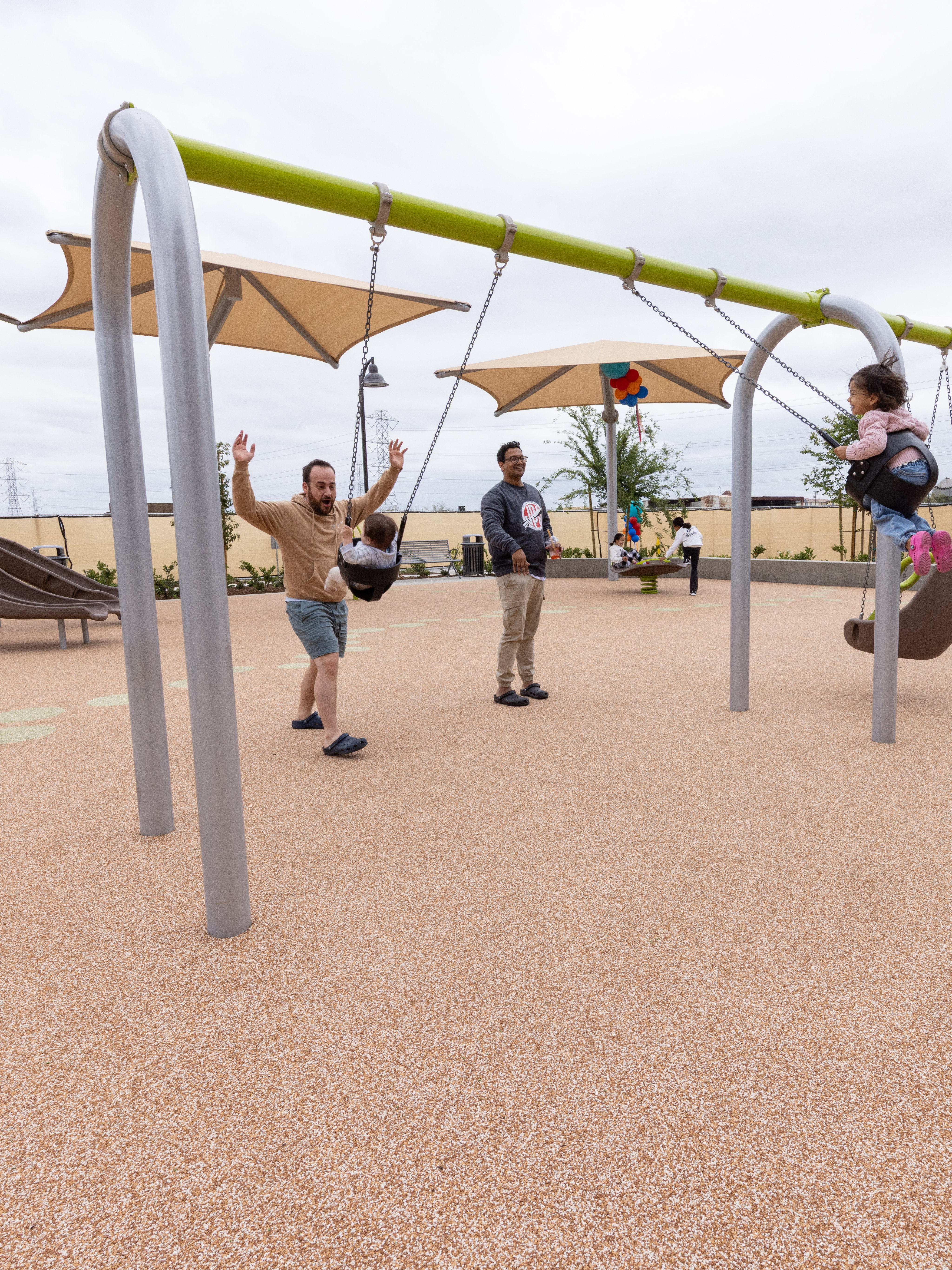A modern outdoor playground with various colorful equipment and structures, surrounded by residential buildings and shaded areas.
