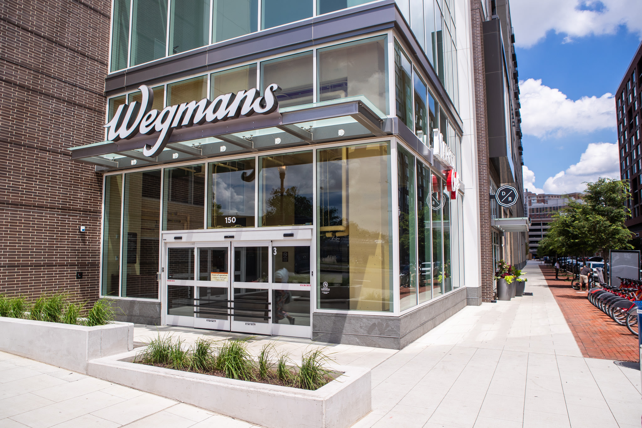 A modern glass-fronted building with the Wegmans logo prominently displayed, surrounded by a paved walkway and landscaping, set against a backdrop of a blue sky with clouds.