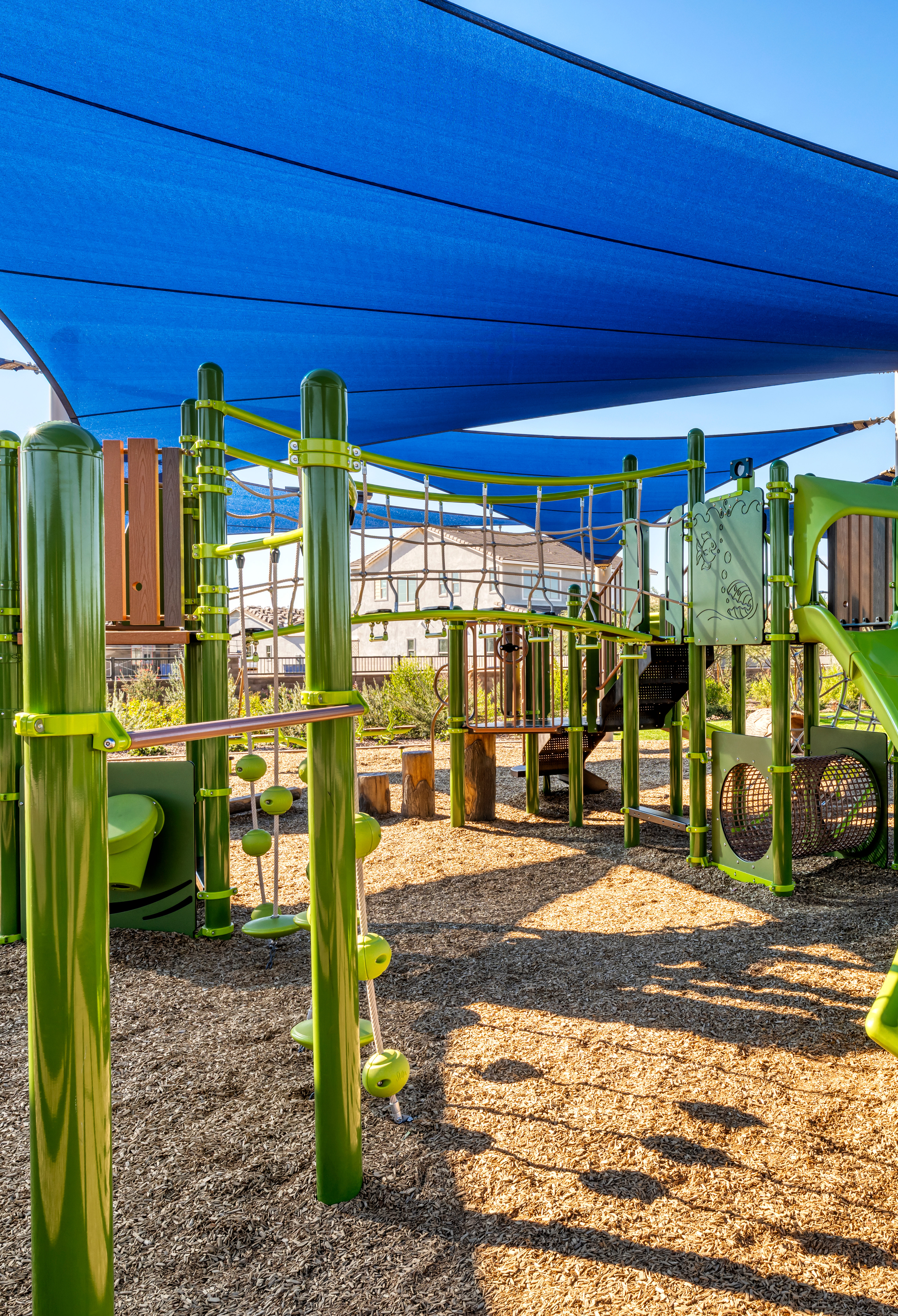 A colorful playground with a blue shade canopy, green and yellow slides, and a paved area surrounded by trees and buildings in the background.