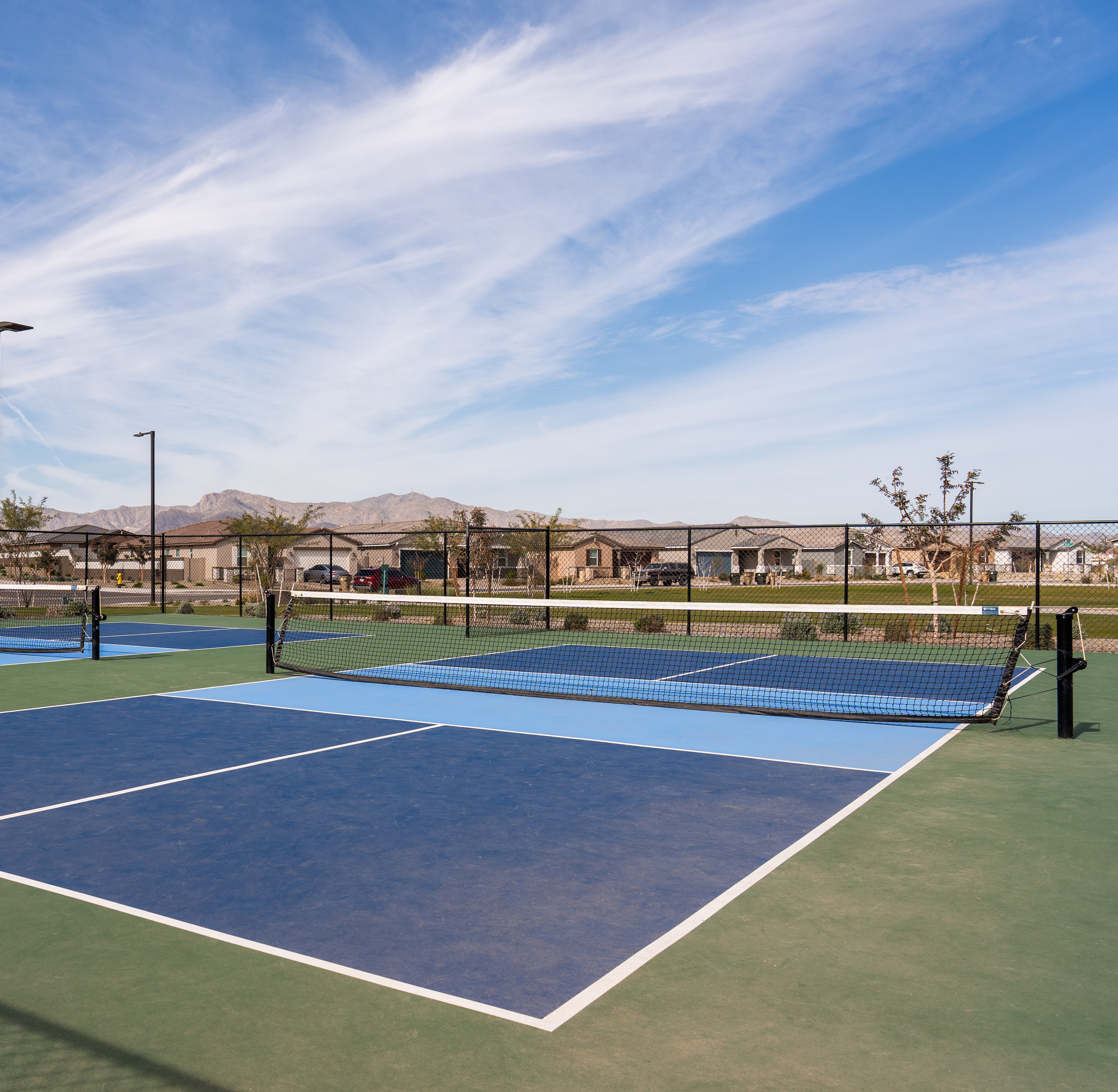 The image shows a tennis court surrounded by residential buildings and a mountainous landscape in the background, with a clear blue sky overhead.