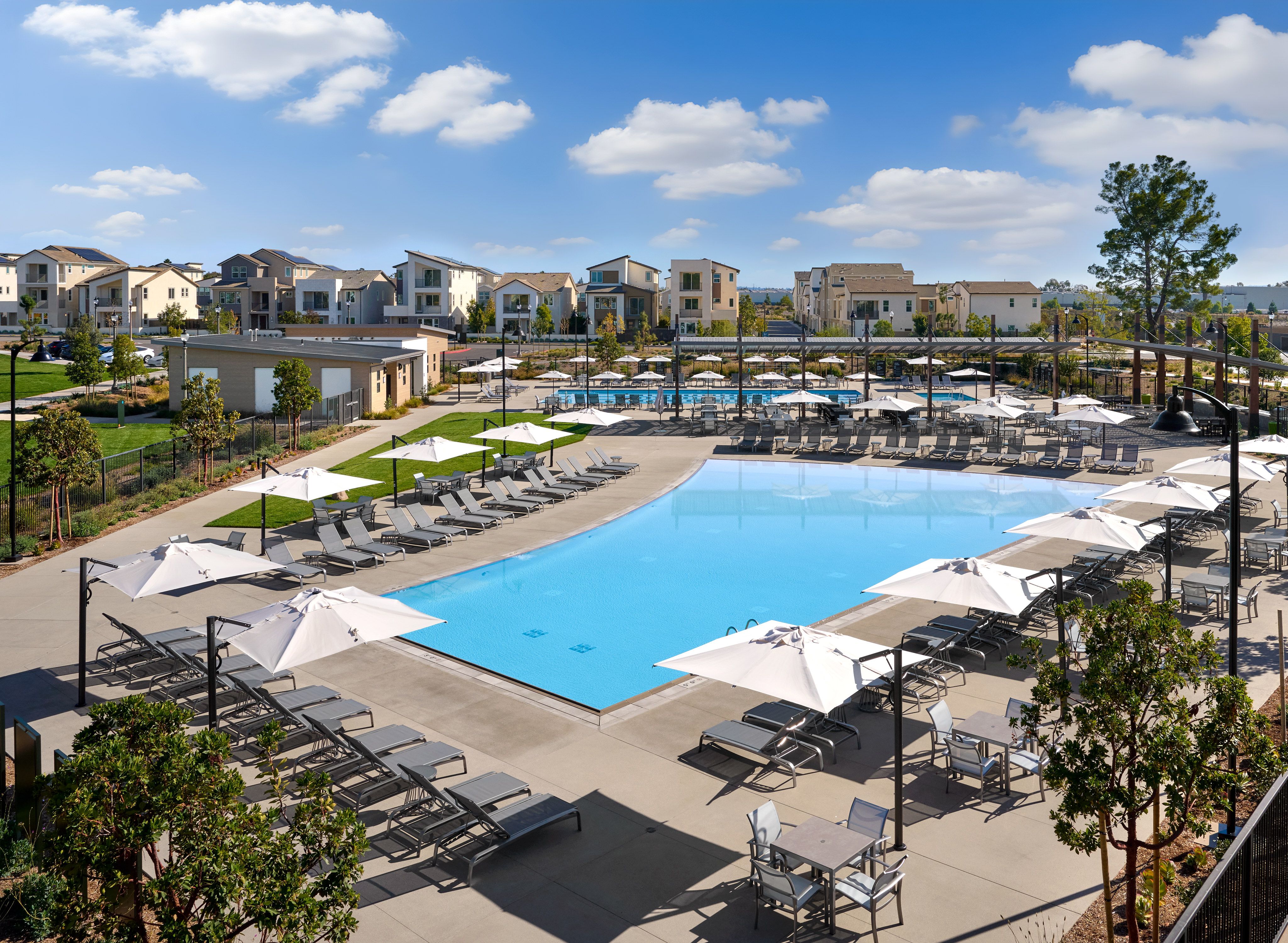 A large outdoor swimming pool surrounded by lounge chairs and umbrellas, with a residential community visible in the background under a blue sky with fluffy white clouds.