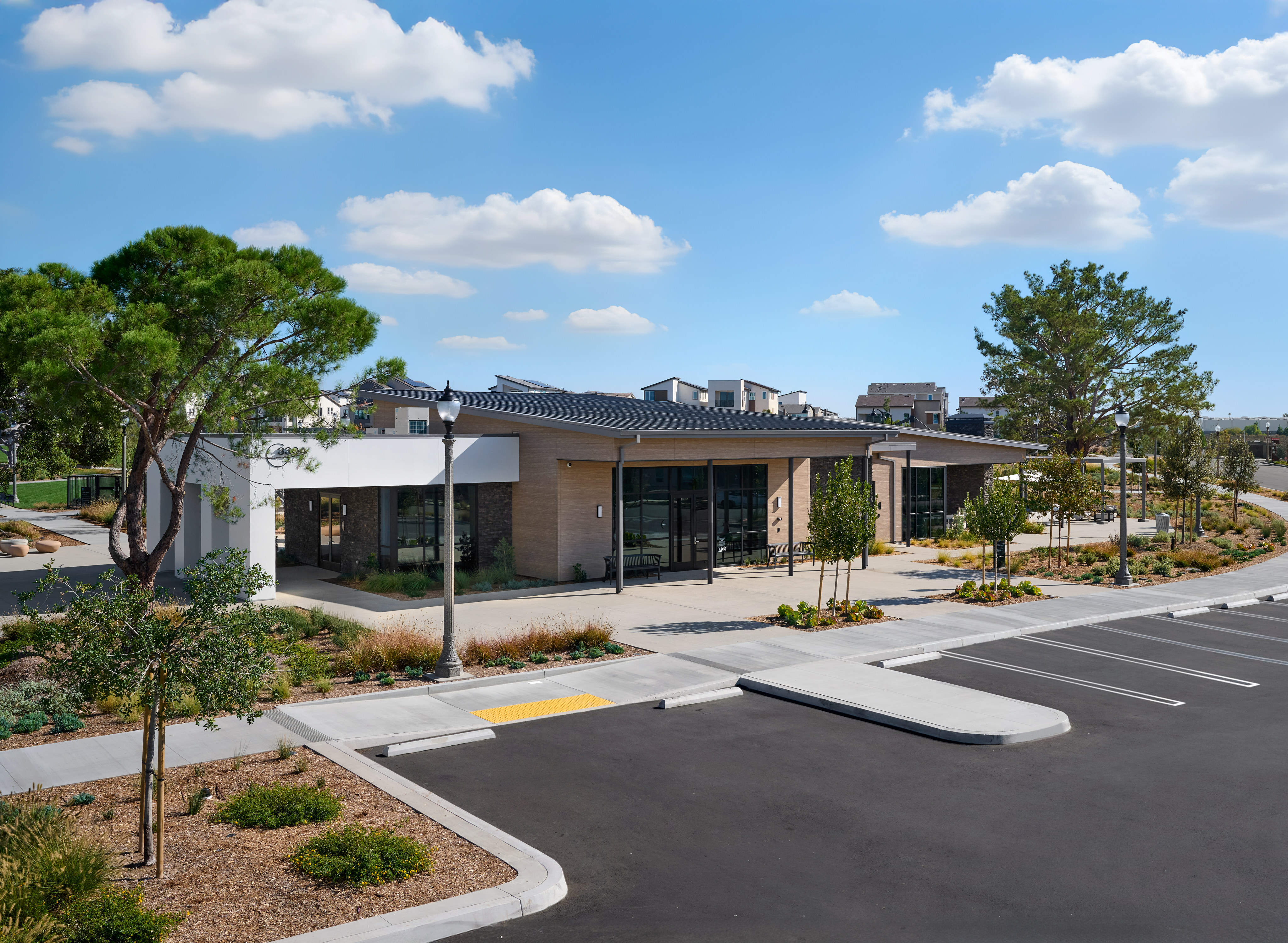 A modern, single-story building with large glass windows and a covered entrance sits in a landscaped area with trees, shrubs, and a paved parking lot in the foreground.