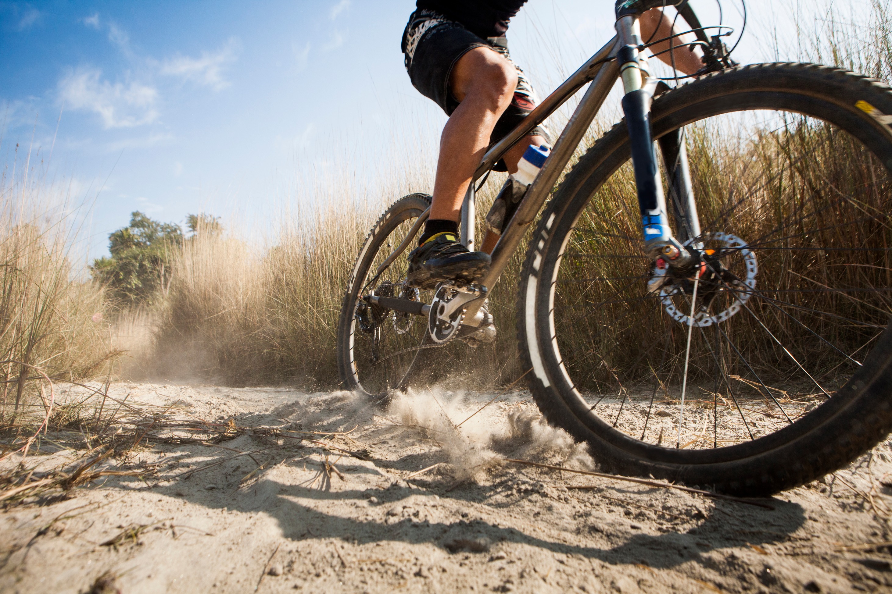 A mountain bike rider navigates a dusty trail surrounded by tall grass and a hazy, sunlit landscape.