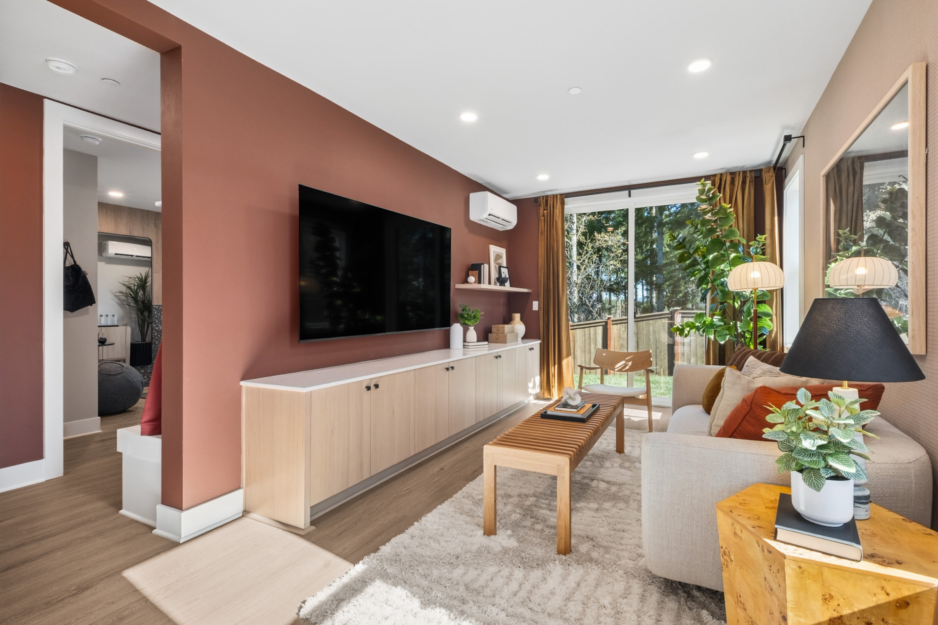 A cozy and modern living room with a large TV mounted on a warm-toned wall, a wooden cabinet below, and a comfortable seating area with a sofa and a wooden bench. The room is filled with natural light from the large windows, and there are potted plants and decorative elements that add to the inviting atmosphere.