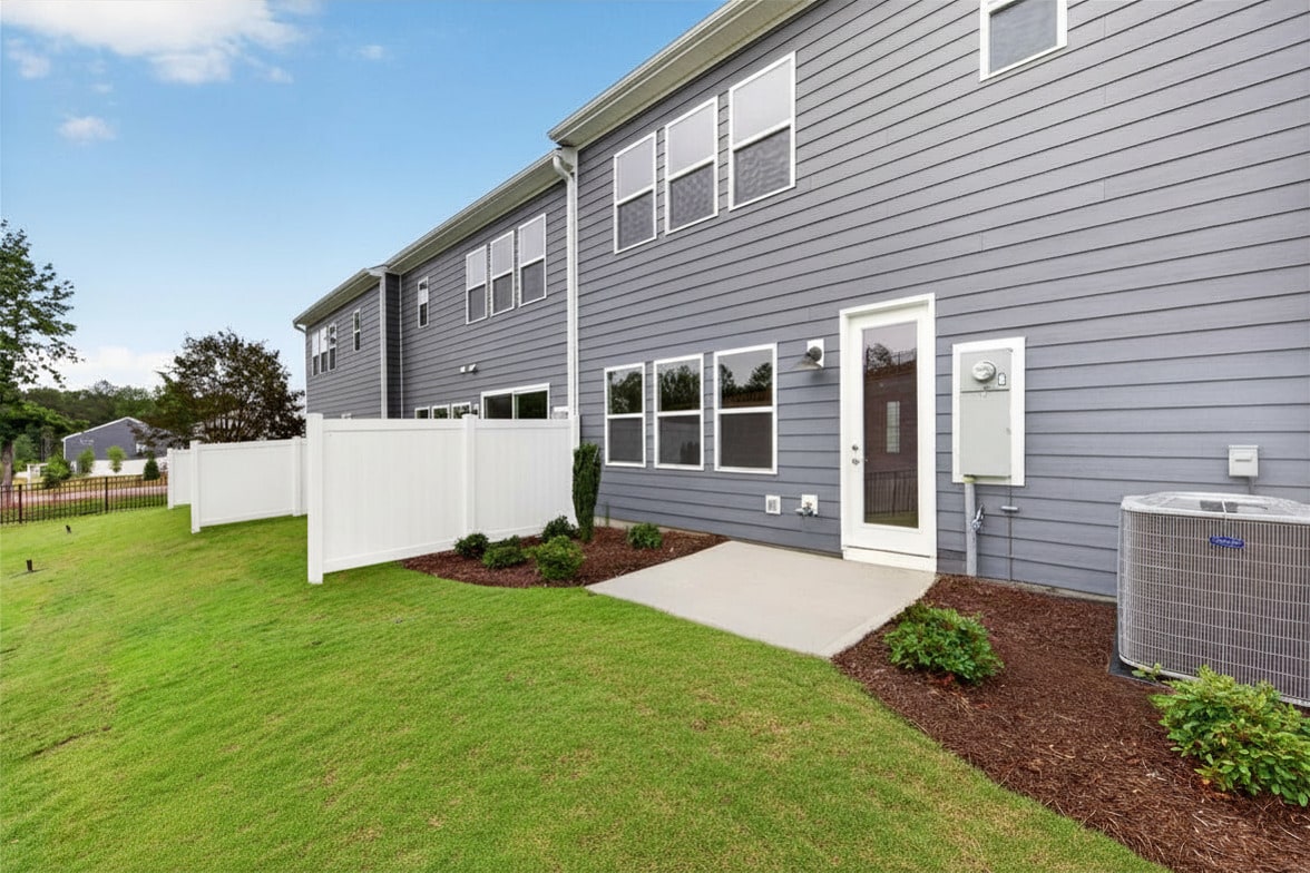 A neat green lawn and white fence are in the foreground, with a row of modern gray townhouses and a blue sky in the background.