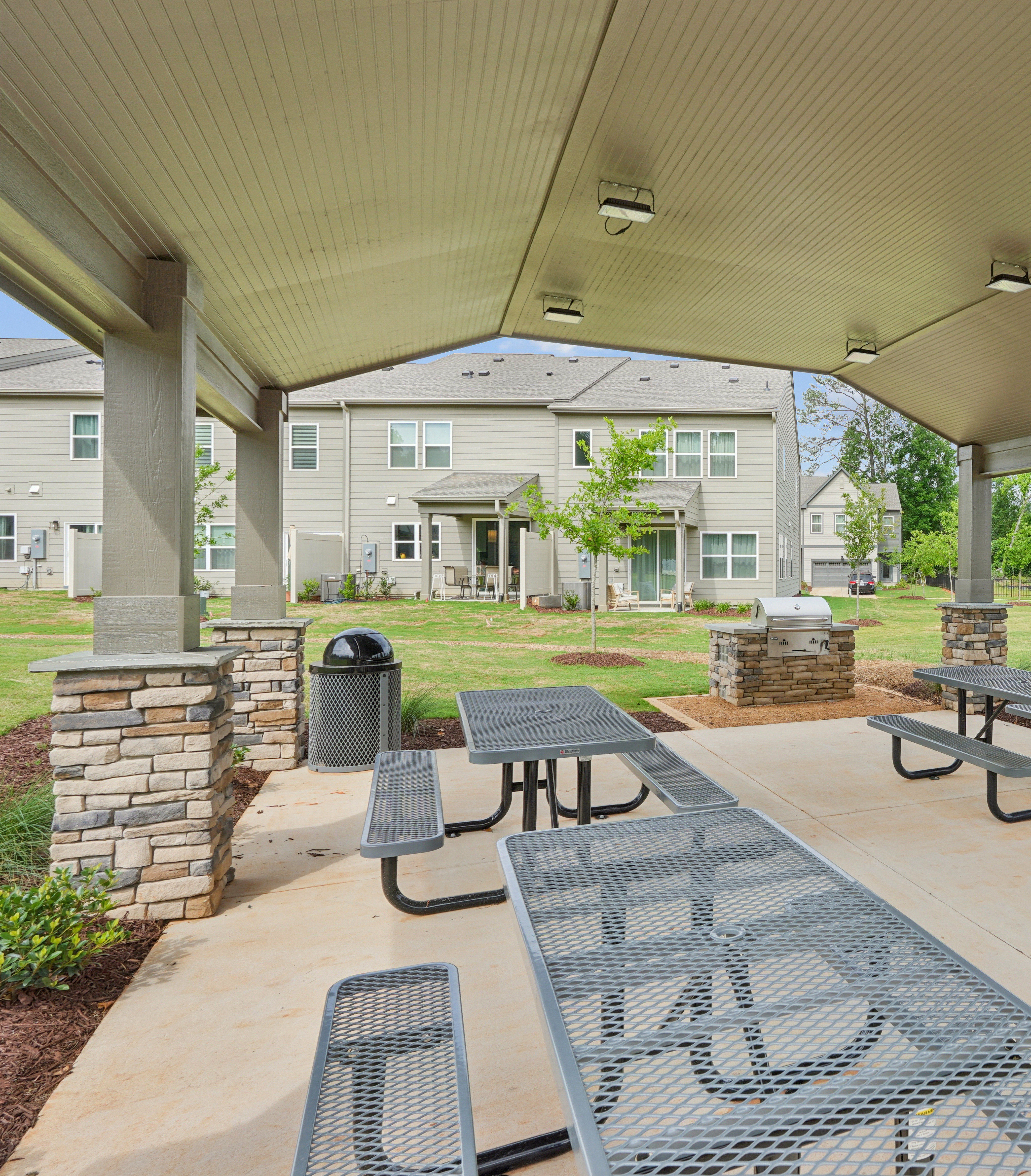Picnic tables and a grill are set up under a covered pavilion in the foreground, with apartment buildings and a grassy field in the background.