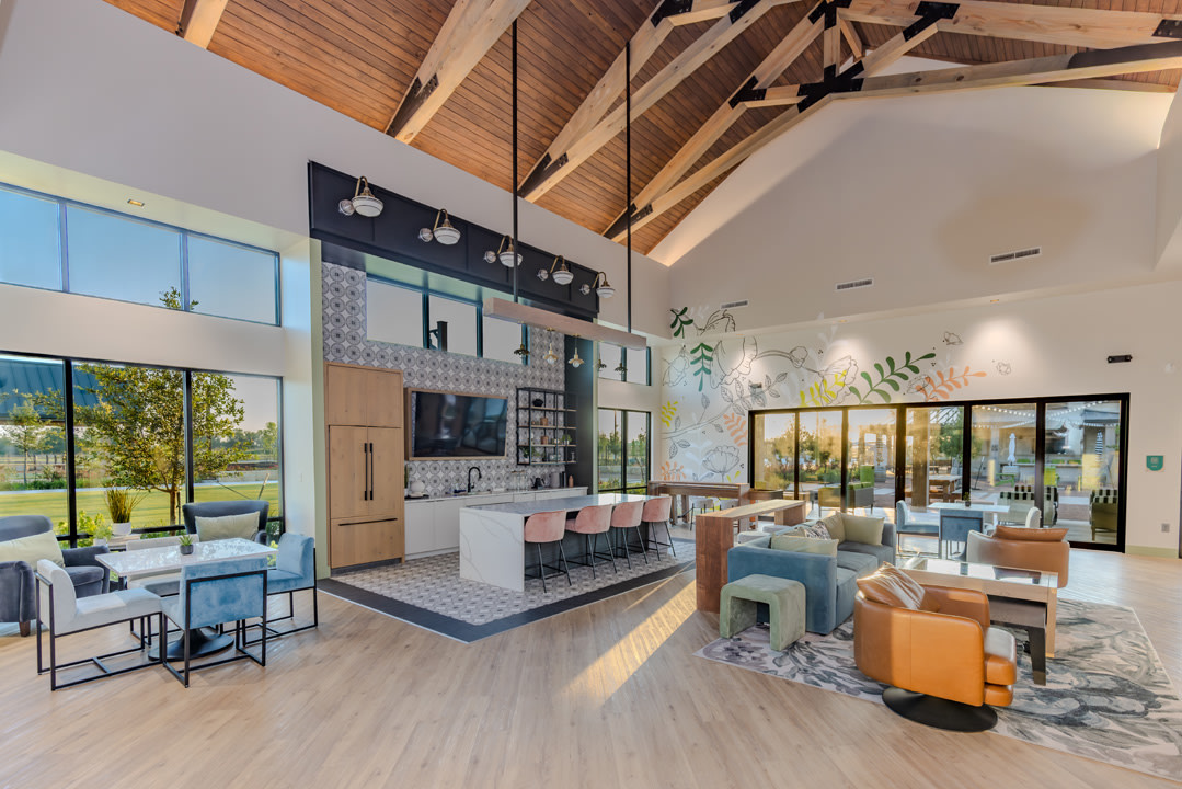 A modern lounge area with a kitchen island and seating in the foreground, and large windows overlooking a sunny outdoor space in the background.