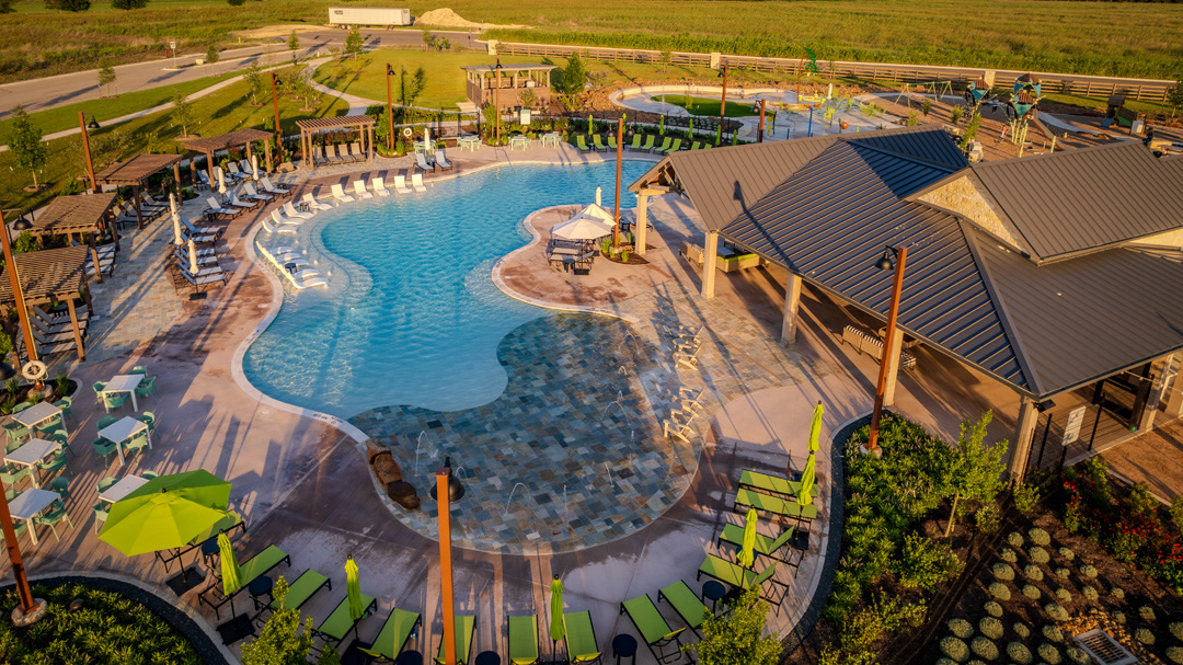 Lush green landscaping and lounge chairs surround a large, freeform swimming pool with covered seating areas, all set against a backdrop of a suburban development and fields.
