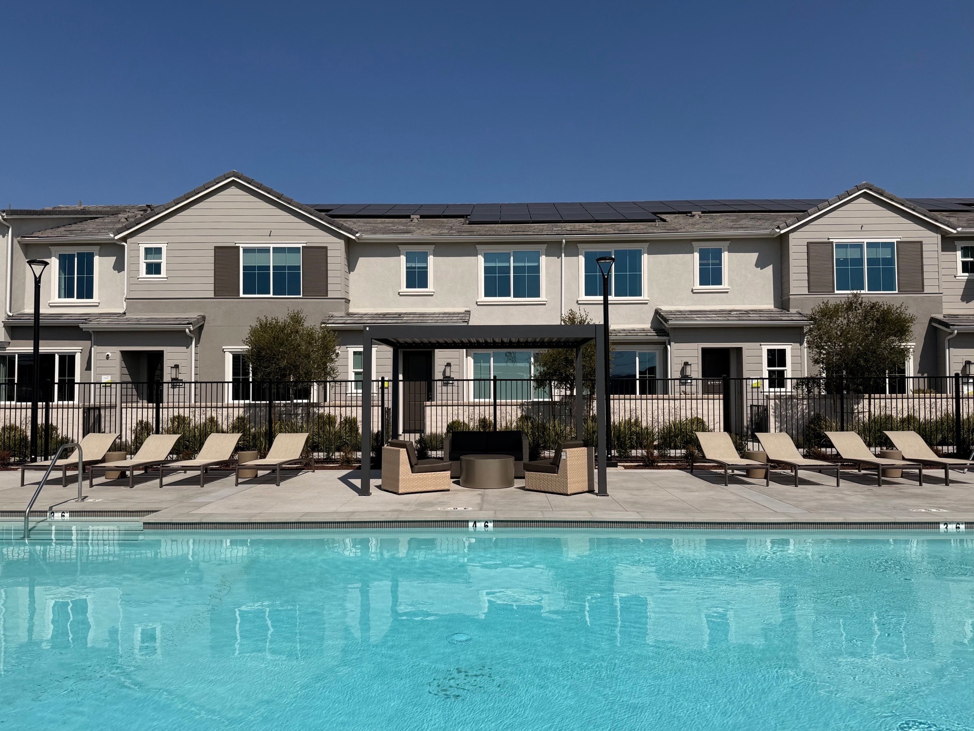 A bright blue swimming pool fills the foreground, with lounge chairs and a fire pit area set against a backdrop of modern townhouses under a clear blue sky.