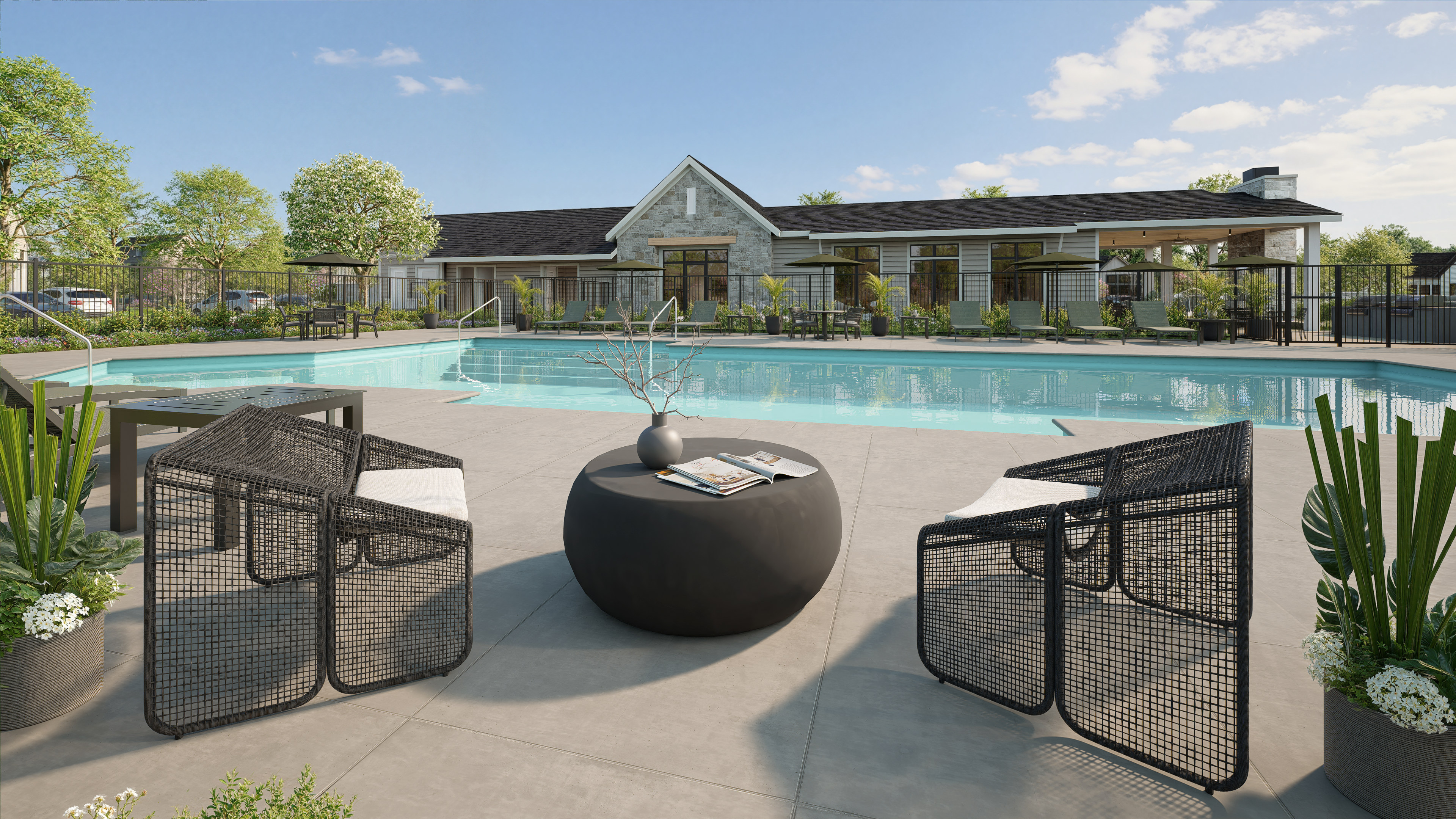 Two modern chairs and a round table with a vase and book sit on a patio in front of a large swimming pool, with a stone-clad building and trees in the background.