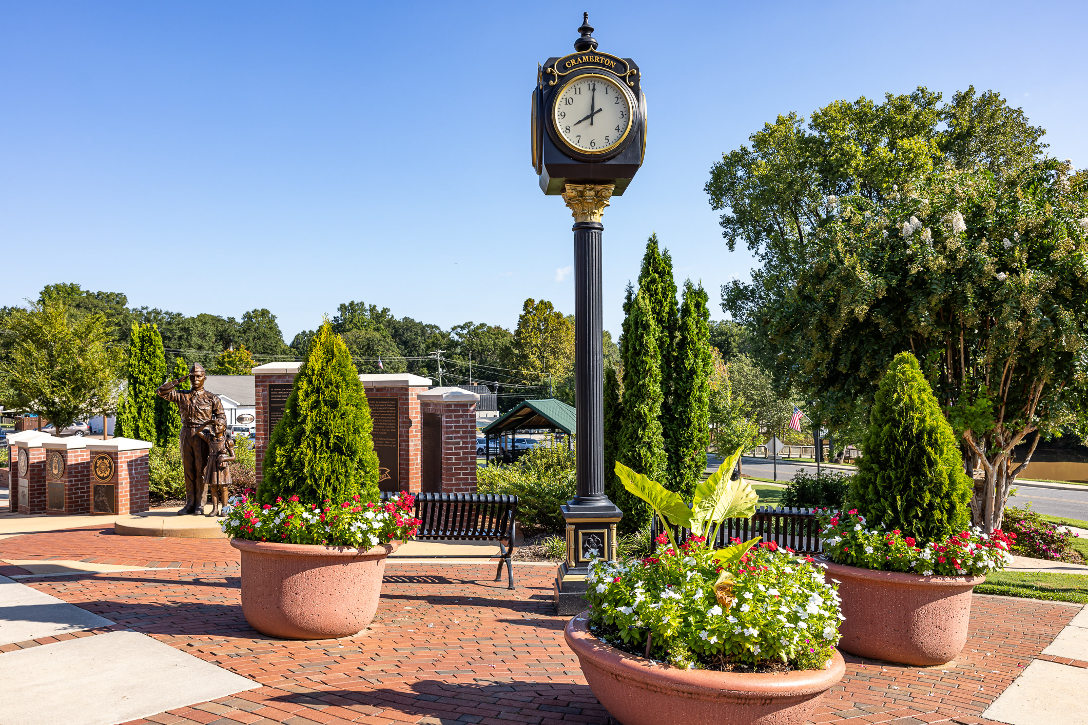 In the foreground, large planters overflow with colorful flowers, while a decorative clock tower stands tall against a background of trees and buildings under a clear blue sky.