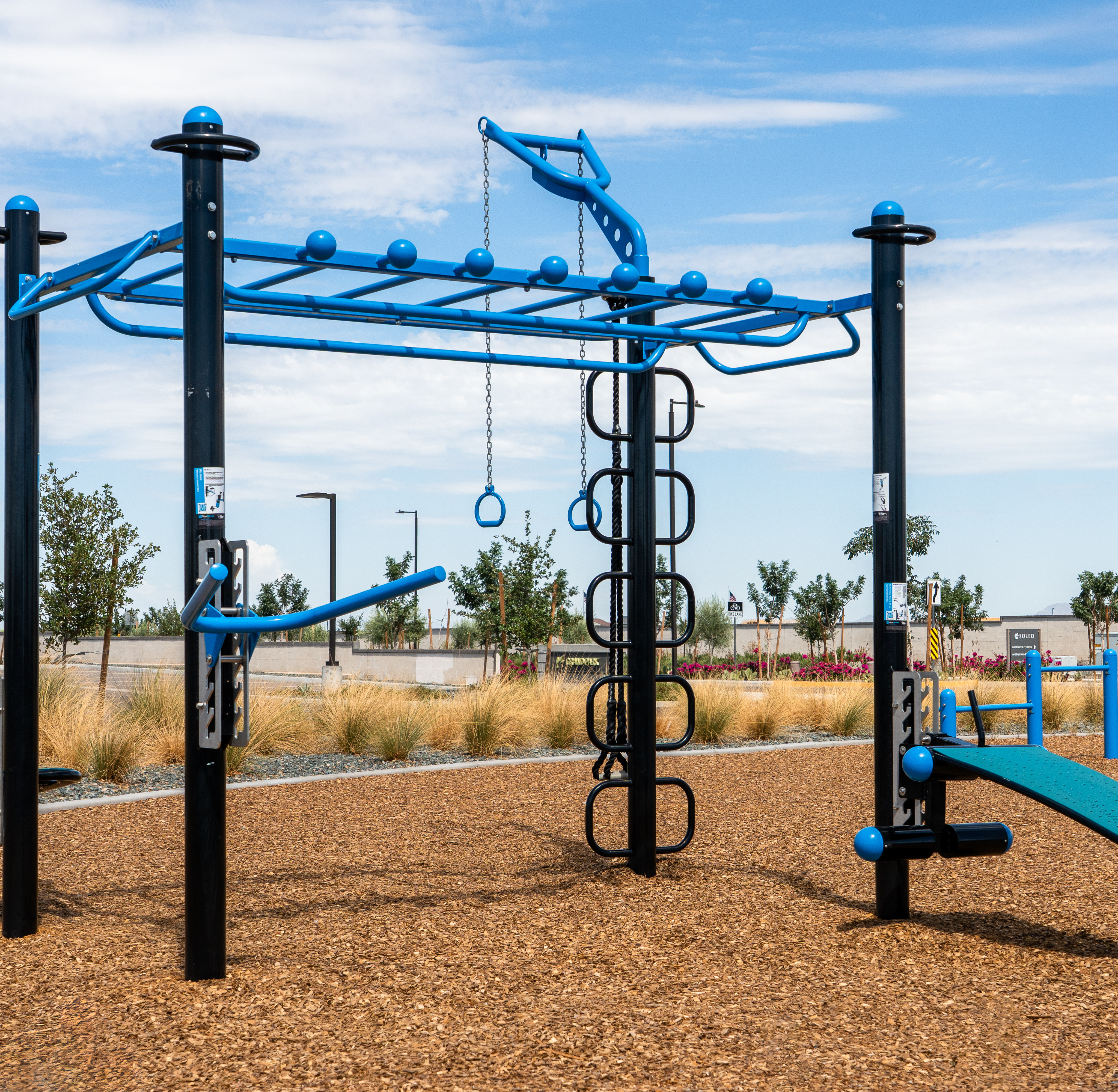 A blue and black outdoor fitness rig stands on a woodchip surface under a bright blue sky with scattered clouds.