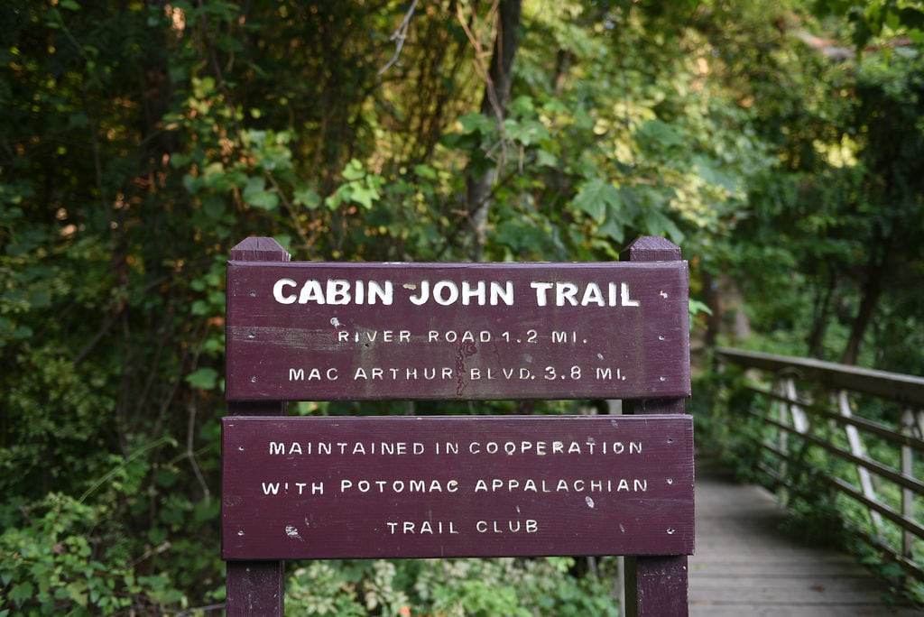 A purple wooden sign for the Cabin John Trail stands in the foreground against a background of lush green foliage and a wooden walkway.