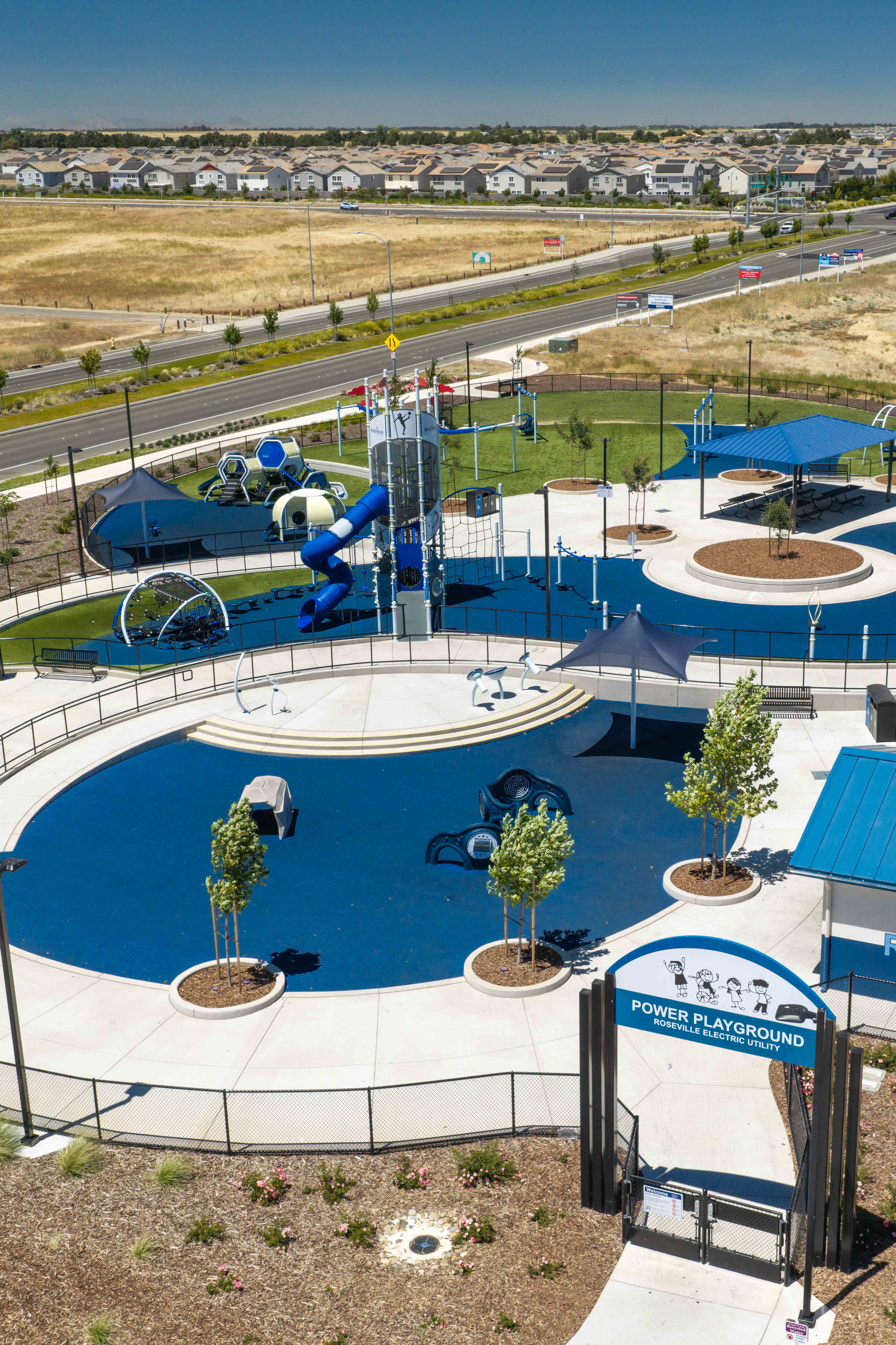 A modern blue and green playground with a restroom building sits in the foreground, while a residential neighborhood and open fields stretch across the background.
