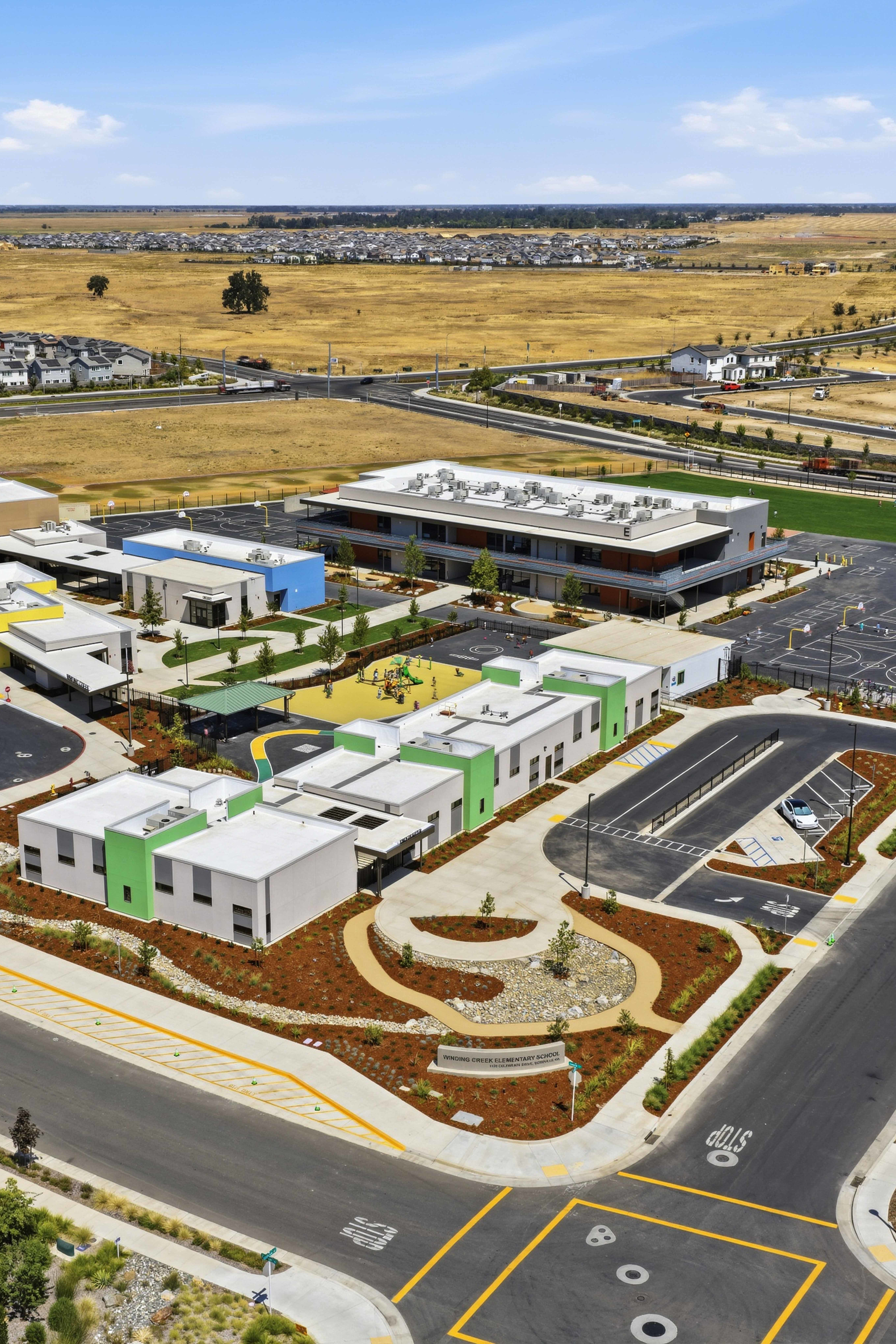 A modern school complex with white and green buildings and parking lots sits in the foreground, while a residential neighborhood and open fields stretch toward the horizon under a blue sky.