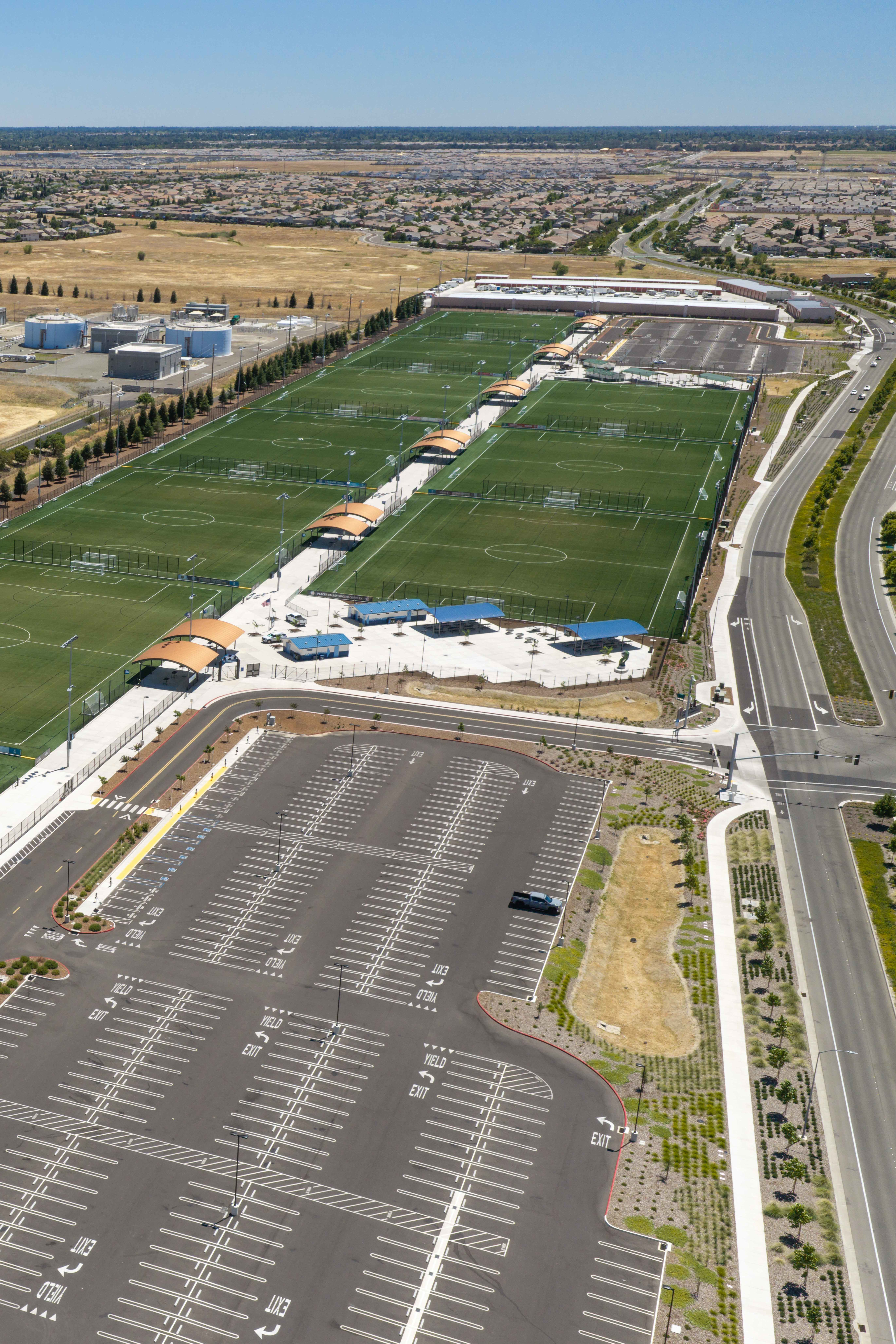 A large parking lot and multiple green soccer fields occupy the foreground, while an industrial area and a sprawling suburban landscape stretch across the background.