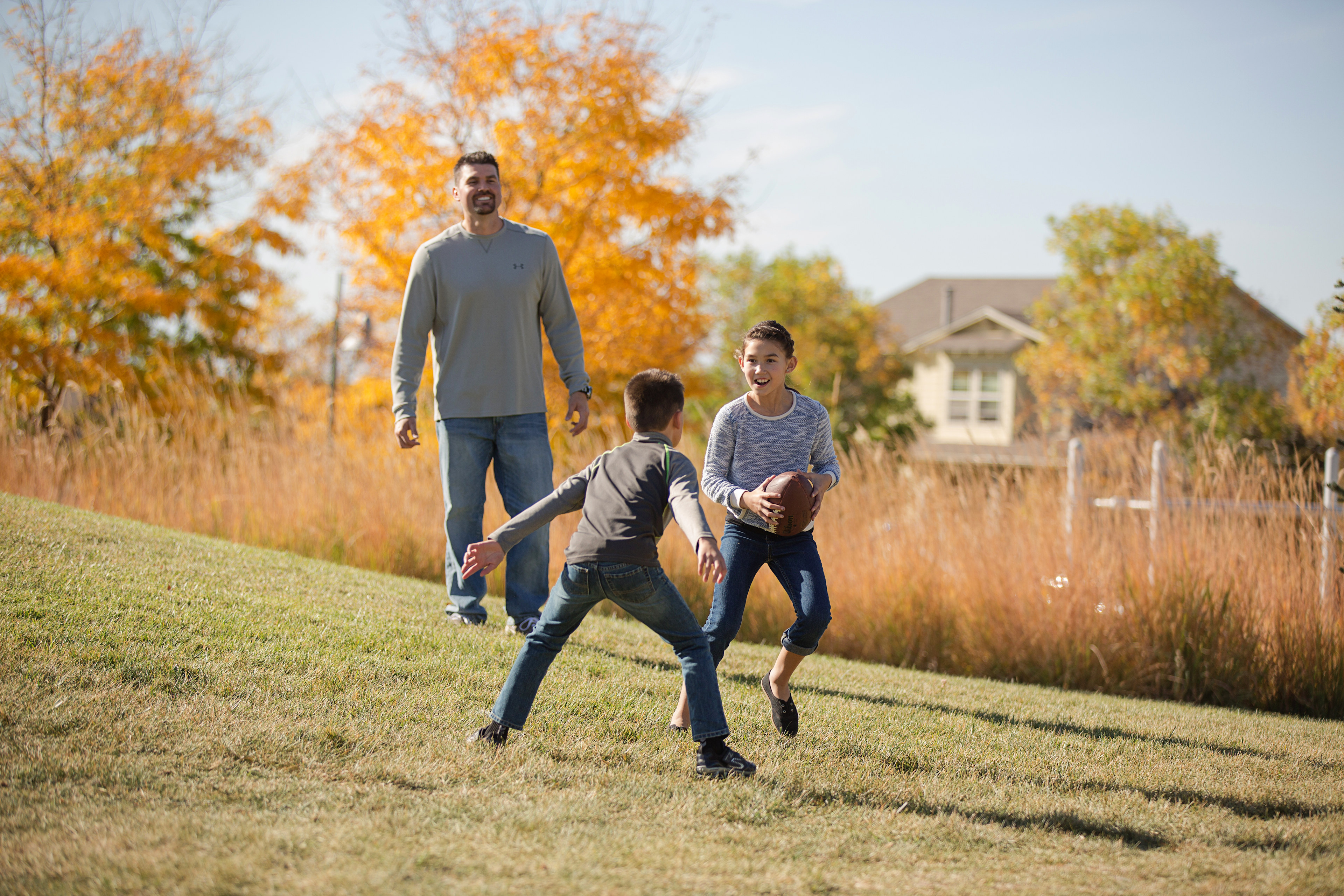 A family of three walking together on a grassy path surrounded by vibrant autumn foliage and a residential neighborhood in the background.